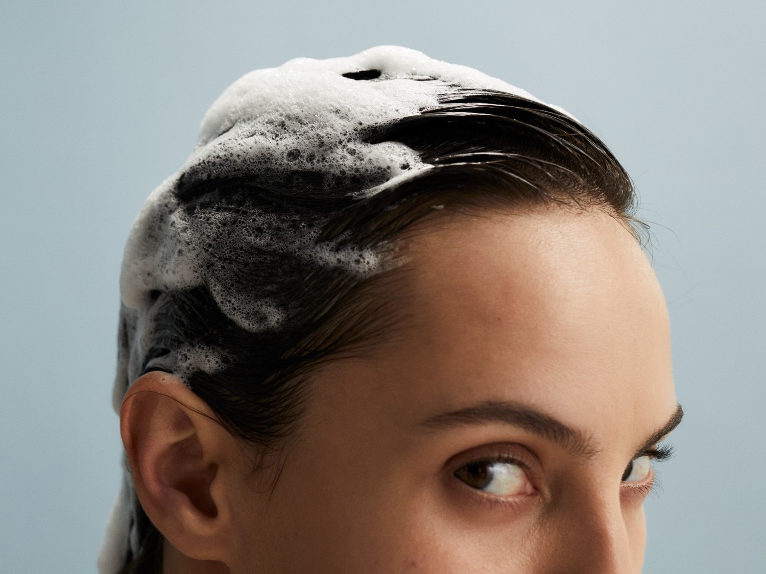 Nahaufnahme einer Frau mit dunklem, nassem Haar, das mit weißem, schaumigem Shampoo bedeckt ist, vor einem hellblauen Hintergrund.