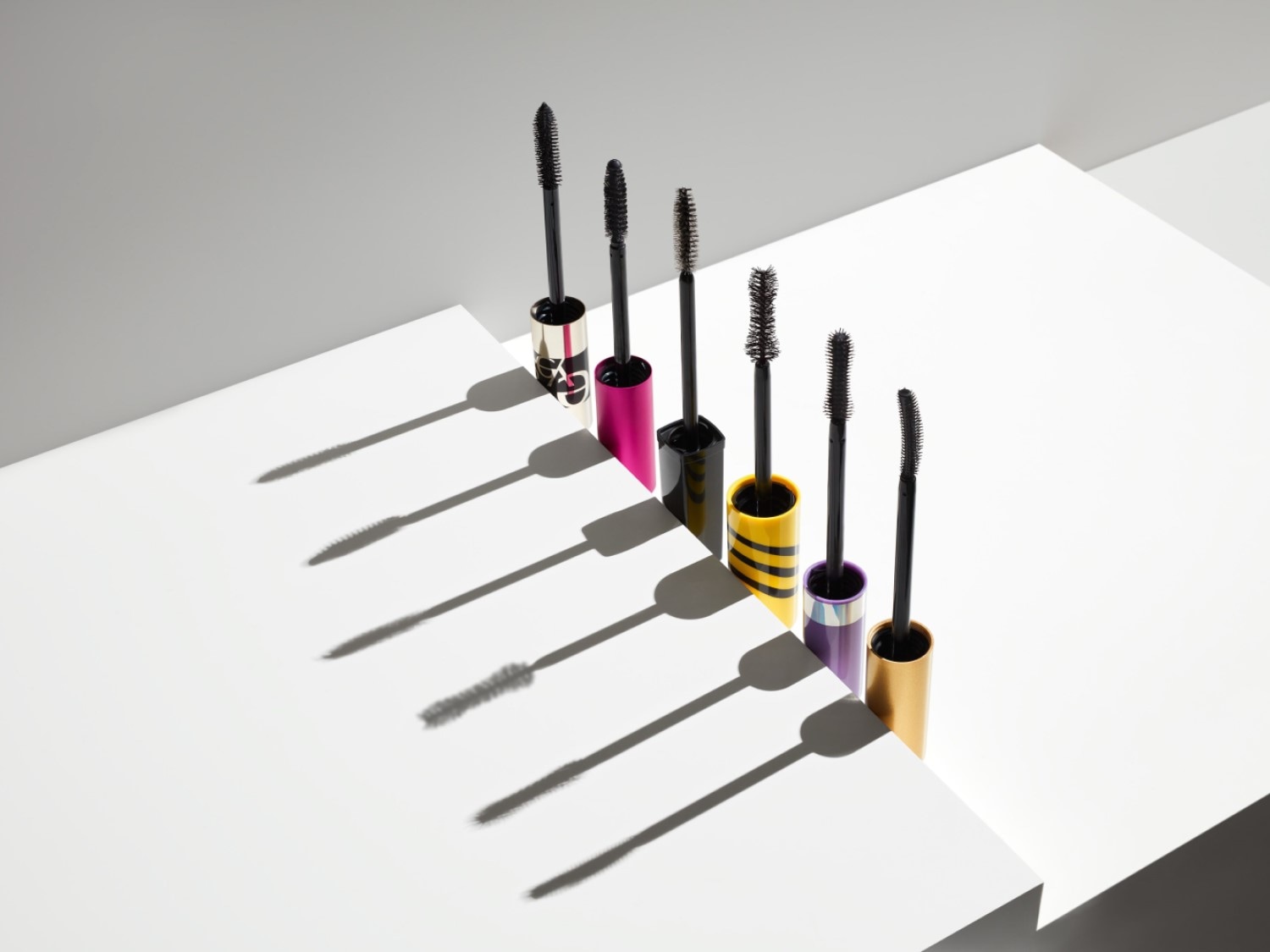 Zes kleurrijke mascara's staan op een witte achtergrond, elk met een zwarte borstel die een lange schaduw werpt.