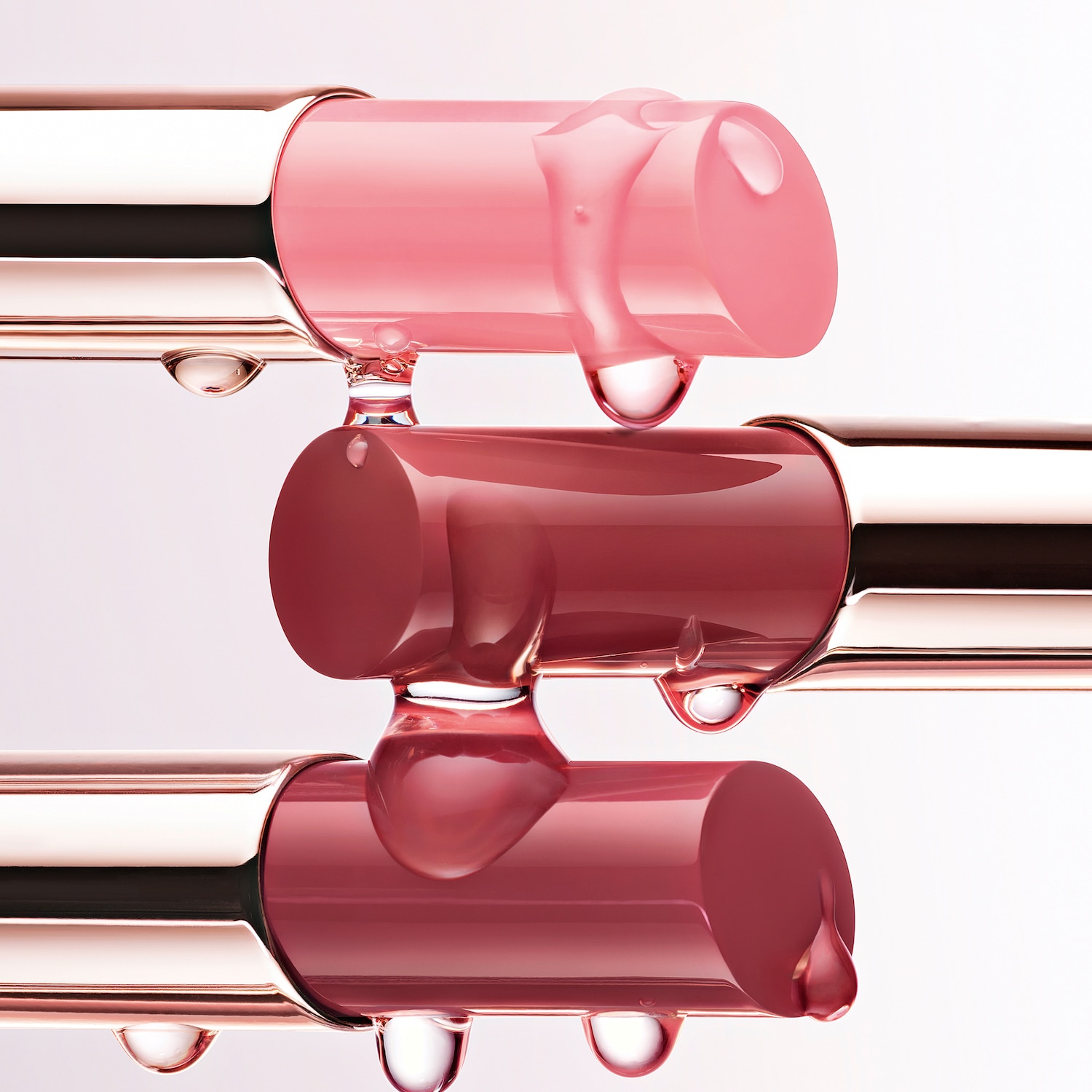 Nahaufnahme von drei übereinander gestapelten Lippenstiften in verschiedenen Rosatönen, die durch transparente Tropfen verbunden sind.