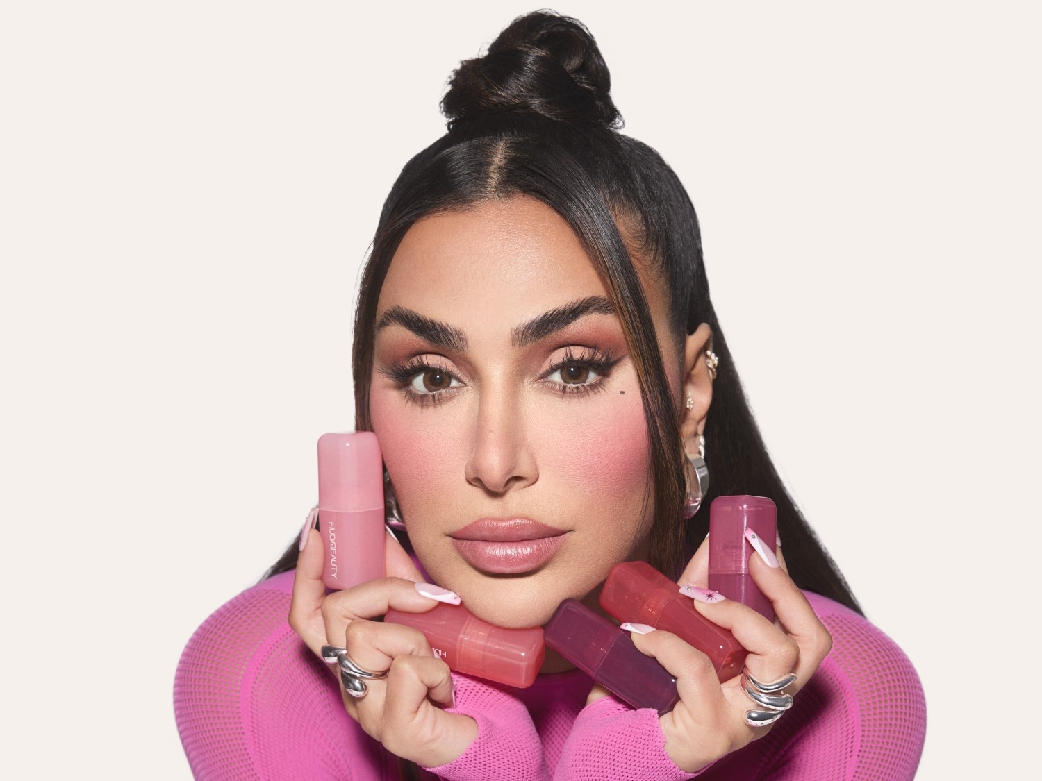 Moteris su rožiniu megztiniu laiko rankose kelis Huda Beauty lūpų dažus su rožiniais dangteliais, pabrėždama jų atspalvius ir tekstūrą.