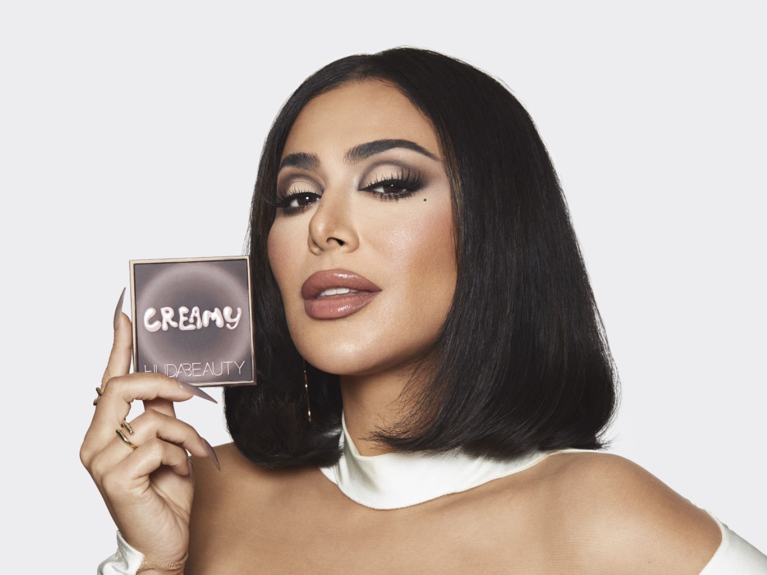 Moteris su ryškiu makiažu laiko Huda Beauty kreminę paletę rudos pakuotėje su auksiniu rėmeliu, ant kurios užrašas „Creamy“.