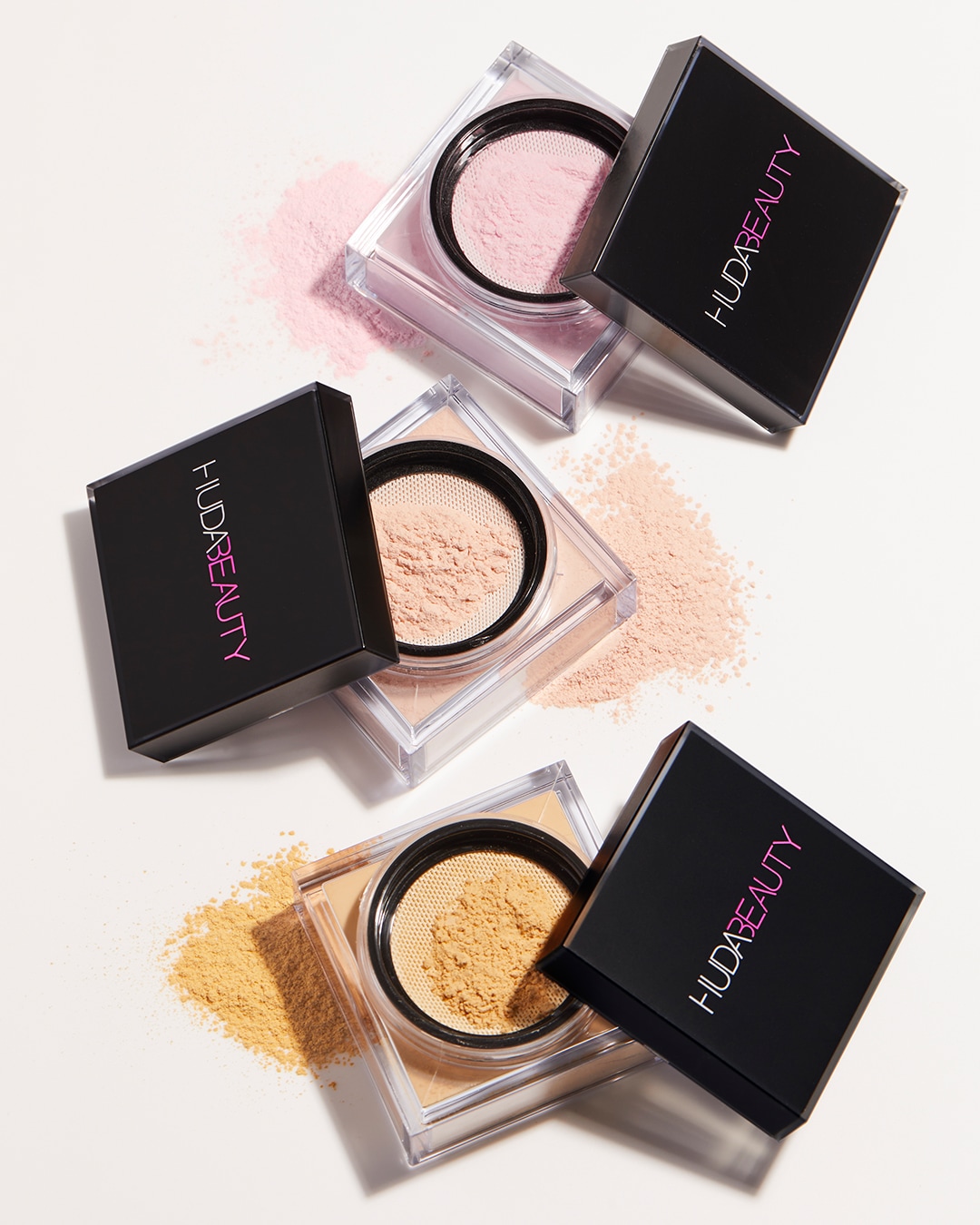 Nuotraukoje matomi trys Huda Beauty biri pudros indai su atidarytais juodais dangteliais, iš kurių byra rožinė, smėlio ir geltona pudra.