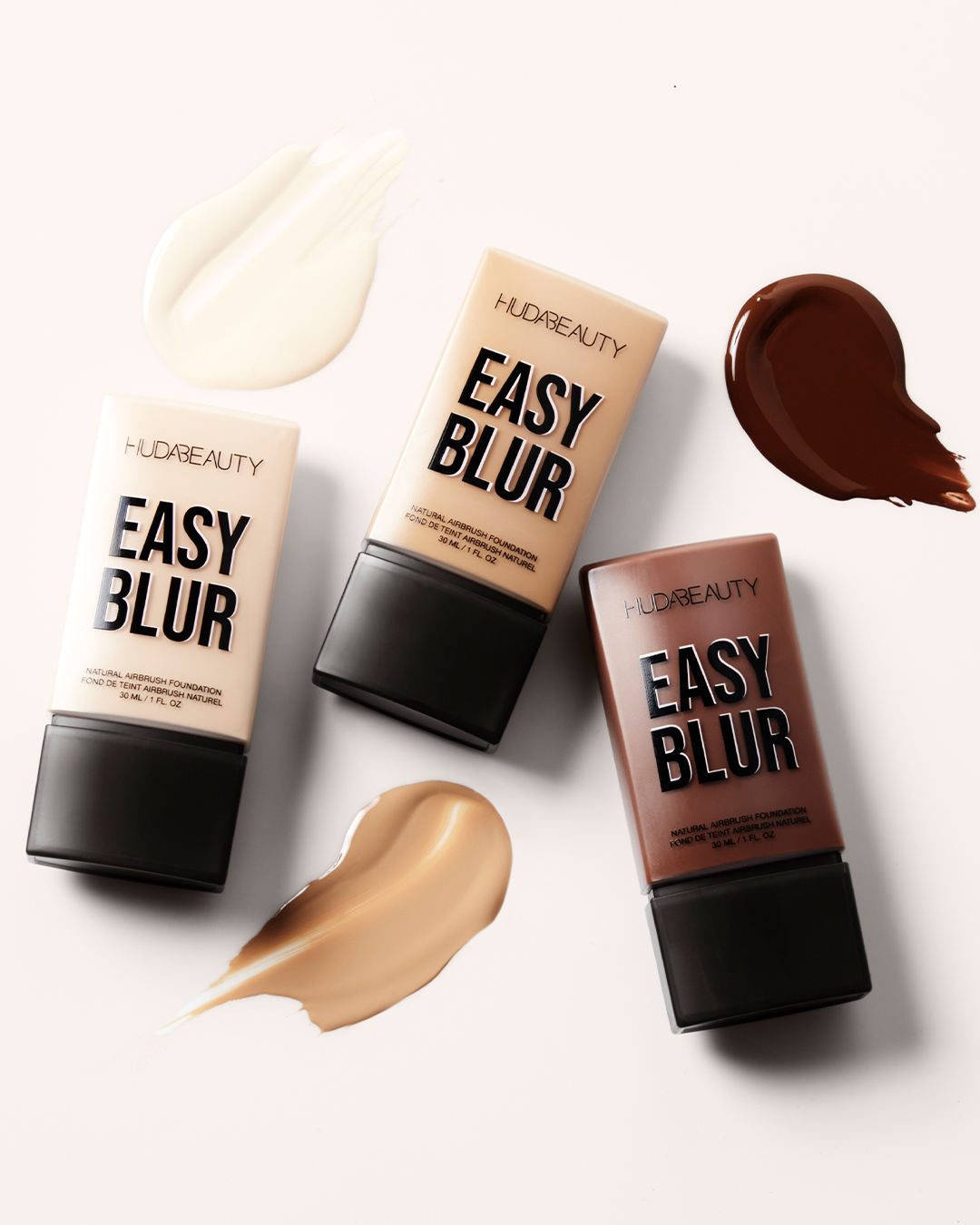 Drei Flaschen Huda Beauty Easy Blur Foundation in verschiedenen Farbtönen mit Swatches, welche die Textur und den jeweiligen Farbton der Foundation zeigen.
