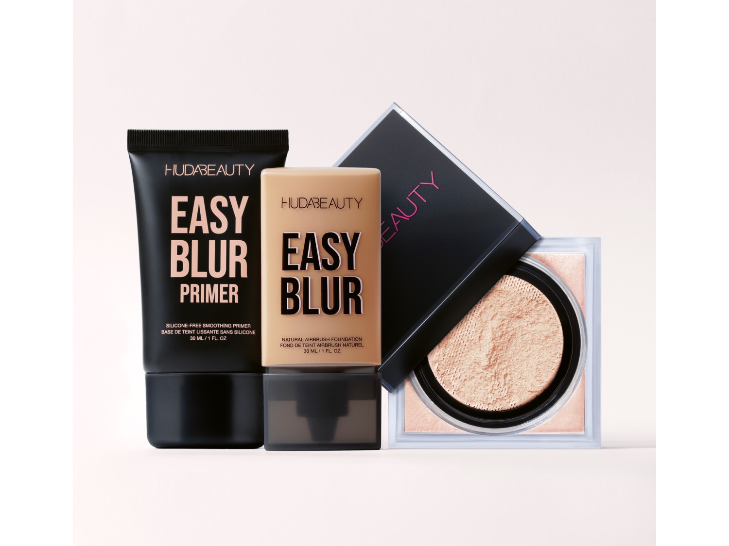 Huda Beauty Easy Blur makiažo pagrindas juodame tūbelėje, šalia smėlio spalvos pagrindo ir birios pudros dėžutėje.