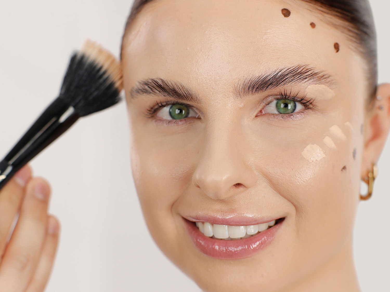 Nahaufnahme einer Frau, die mit einem Pinsel und beiger Creme im Gesicht lächelt, um einen ebenmäßigen Teint zu erzielen.