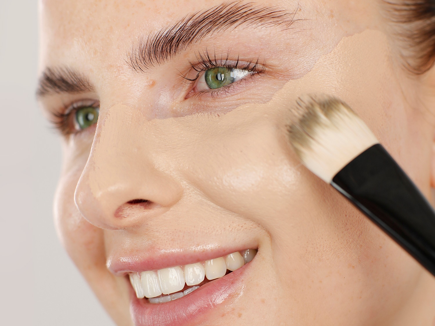 Nahaufnahme einer Frau, die mit einem Pinsel eine helle Grundierung auf ihr Gesicht aufträgt, um einen ebenmäßigen Teint zu erzielen.