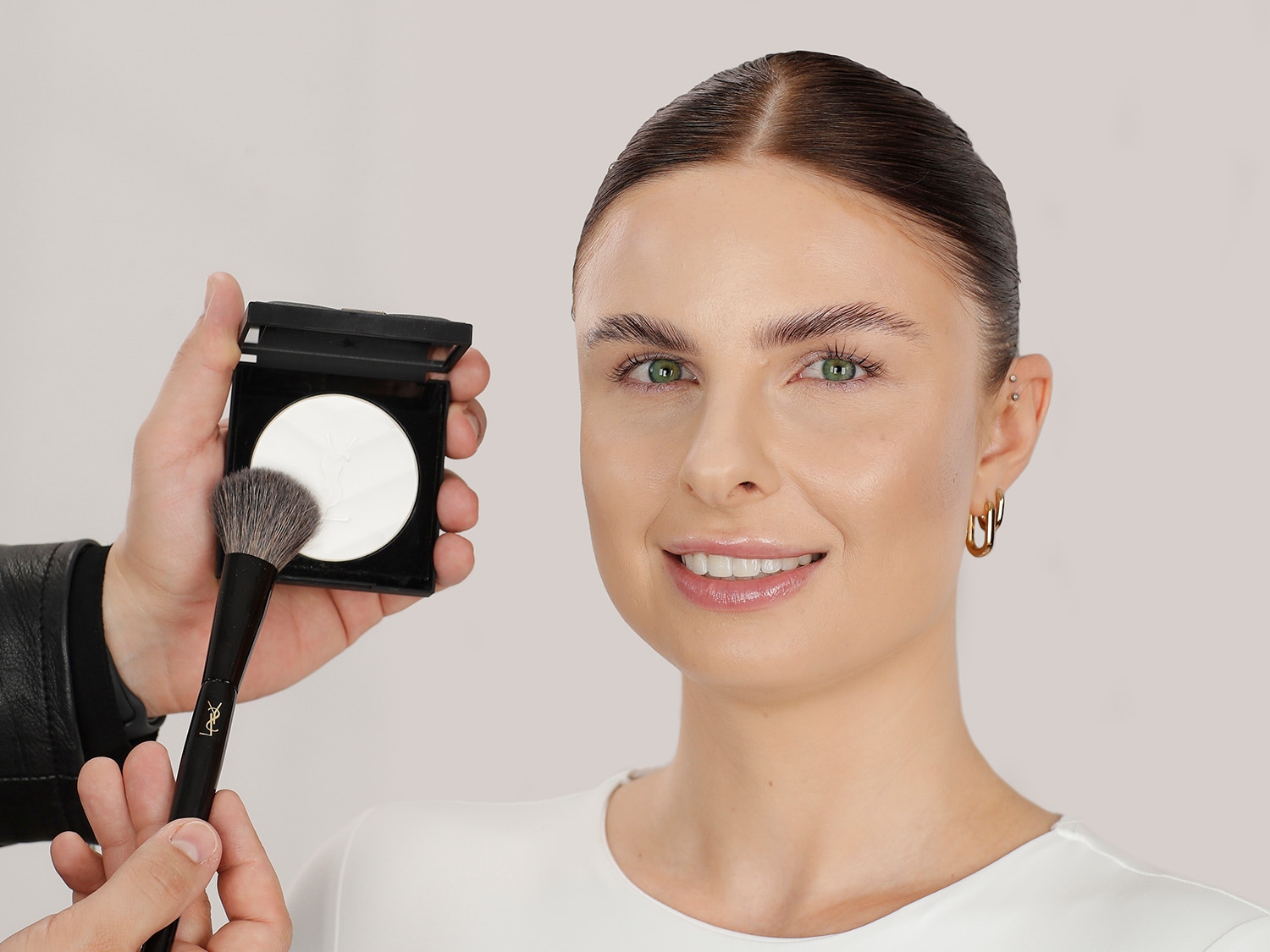 Eine Frau mit strahlendem Lächeln, während ein Make-up-Artist mit einem Pinsel losen, weißen Puder von Yves Saint Laurent auf ihr Gesicht aufträgt.