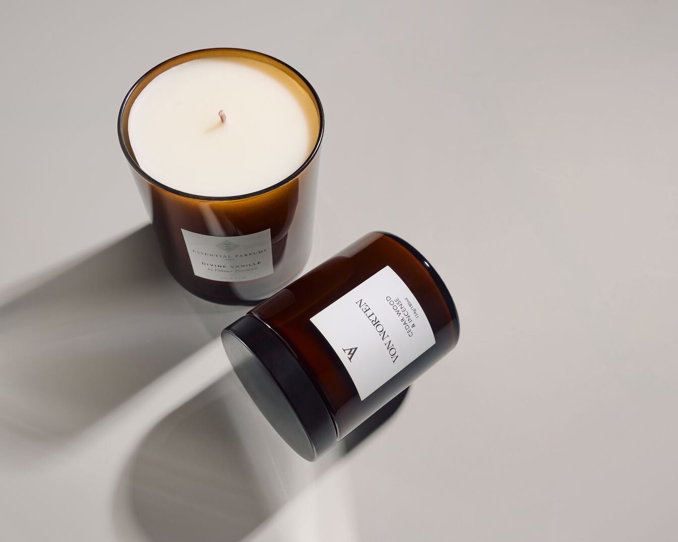 Zwei braune Duftkerzen stehen auf einem hellen Untergrund, die eine aufrecht mit weißem Wachs, die andere liegend, beide mit weißen Etiketten.