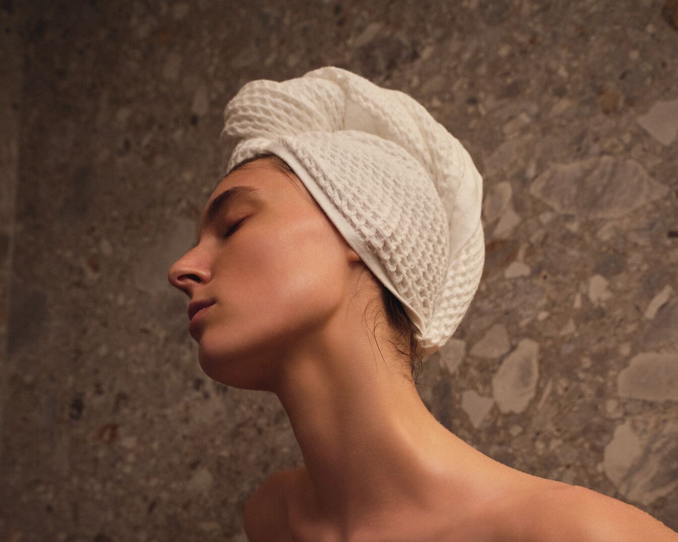 Eine Frau mit geschlossenen Augen und einem weißen, strukturierten Handtuch um den Kopf gewickelt, genießt einen Moment der Ruhe.