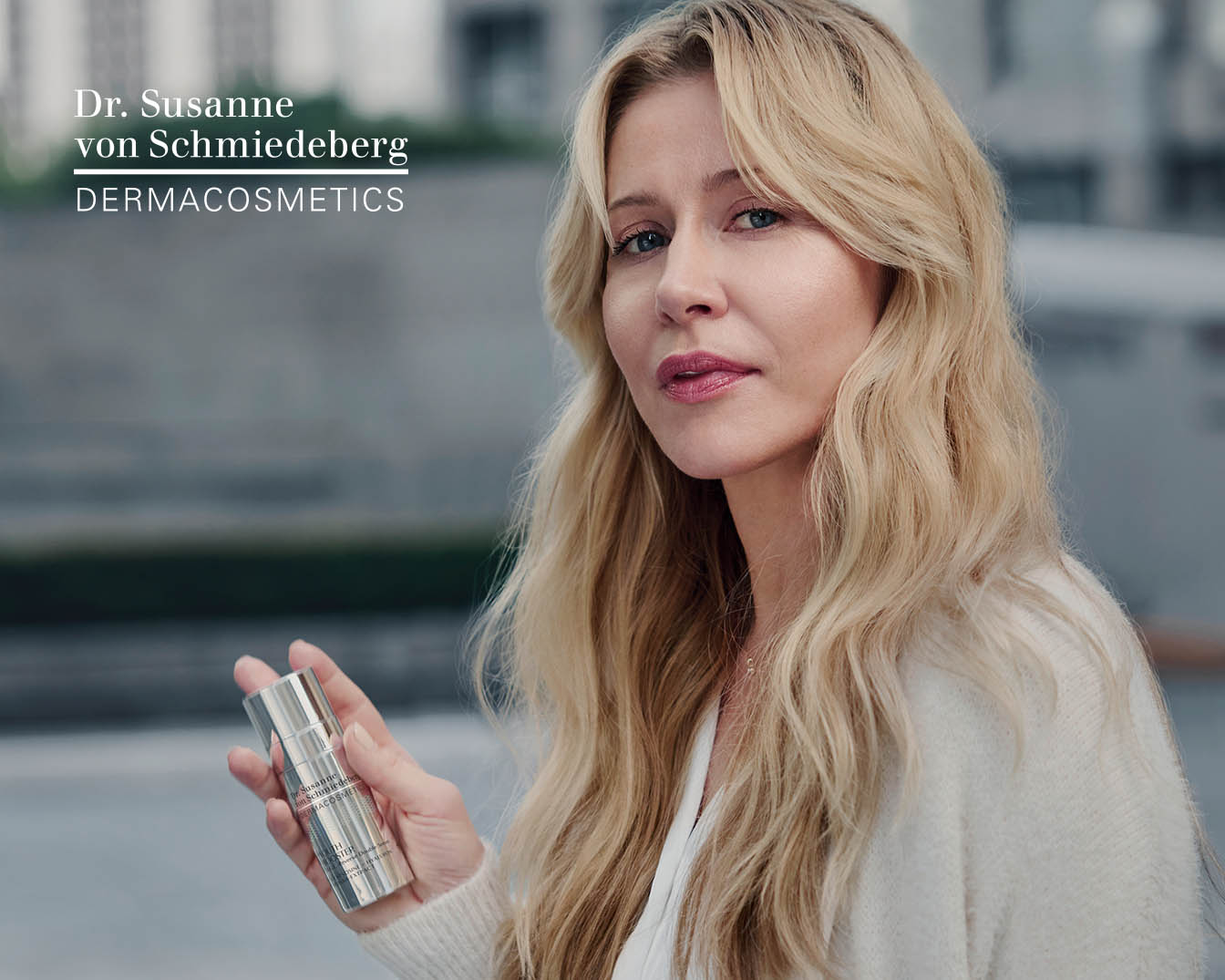 Blondwłosa kobieta prezentuje srebrne opakowanie kosmetyku Dr. Susanne von Schmiedeberg DERMACOSMETICS, trzymając je w dłoni.