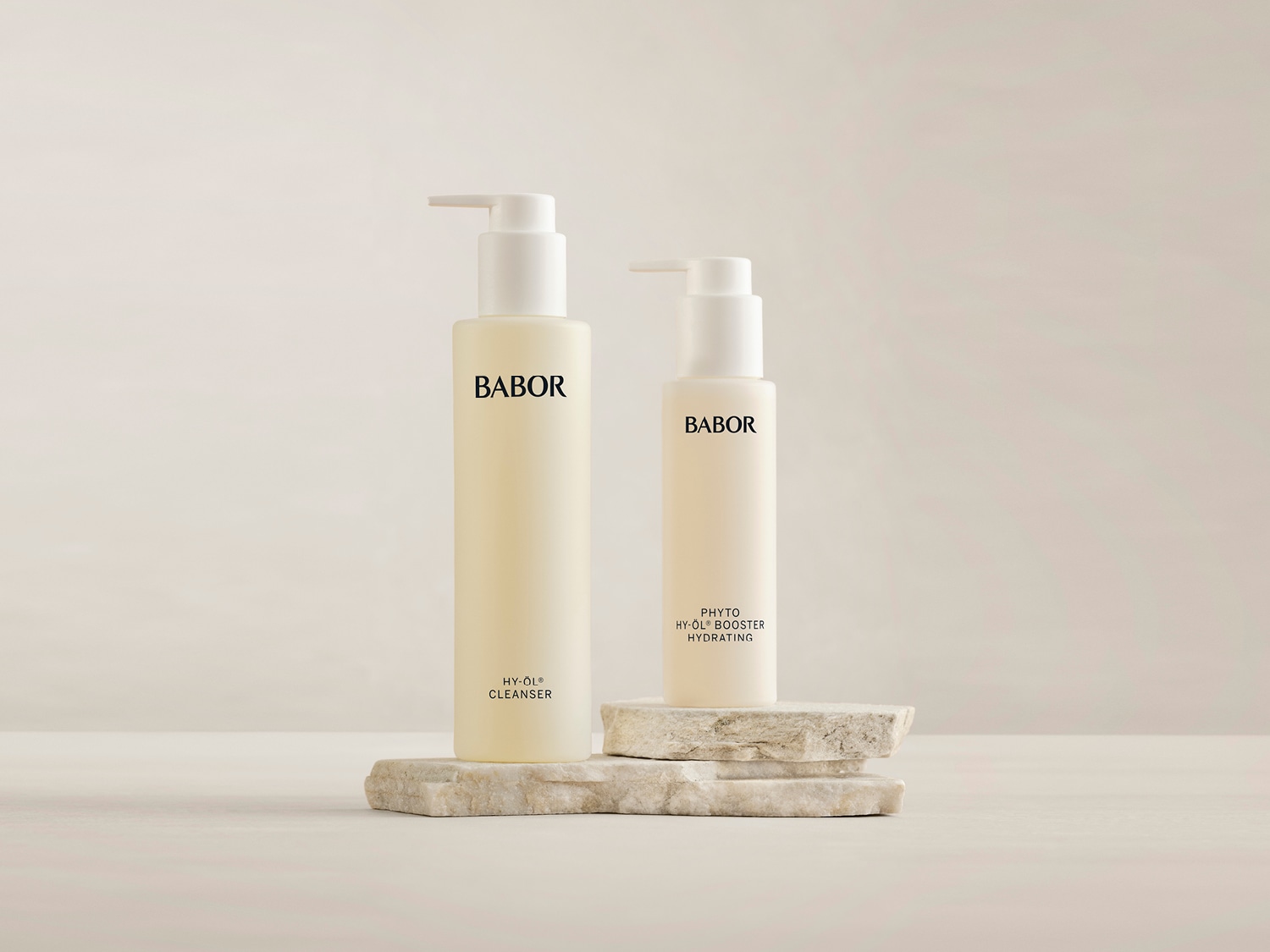 Dwa beżowe flakony Babor: HY-ÖL Cleanser i Phyto HY-ÖL Booster Hydrating, stojące na kamiennych podstawkach, prezentują się minimalistycznie i elegancko.