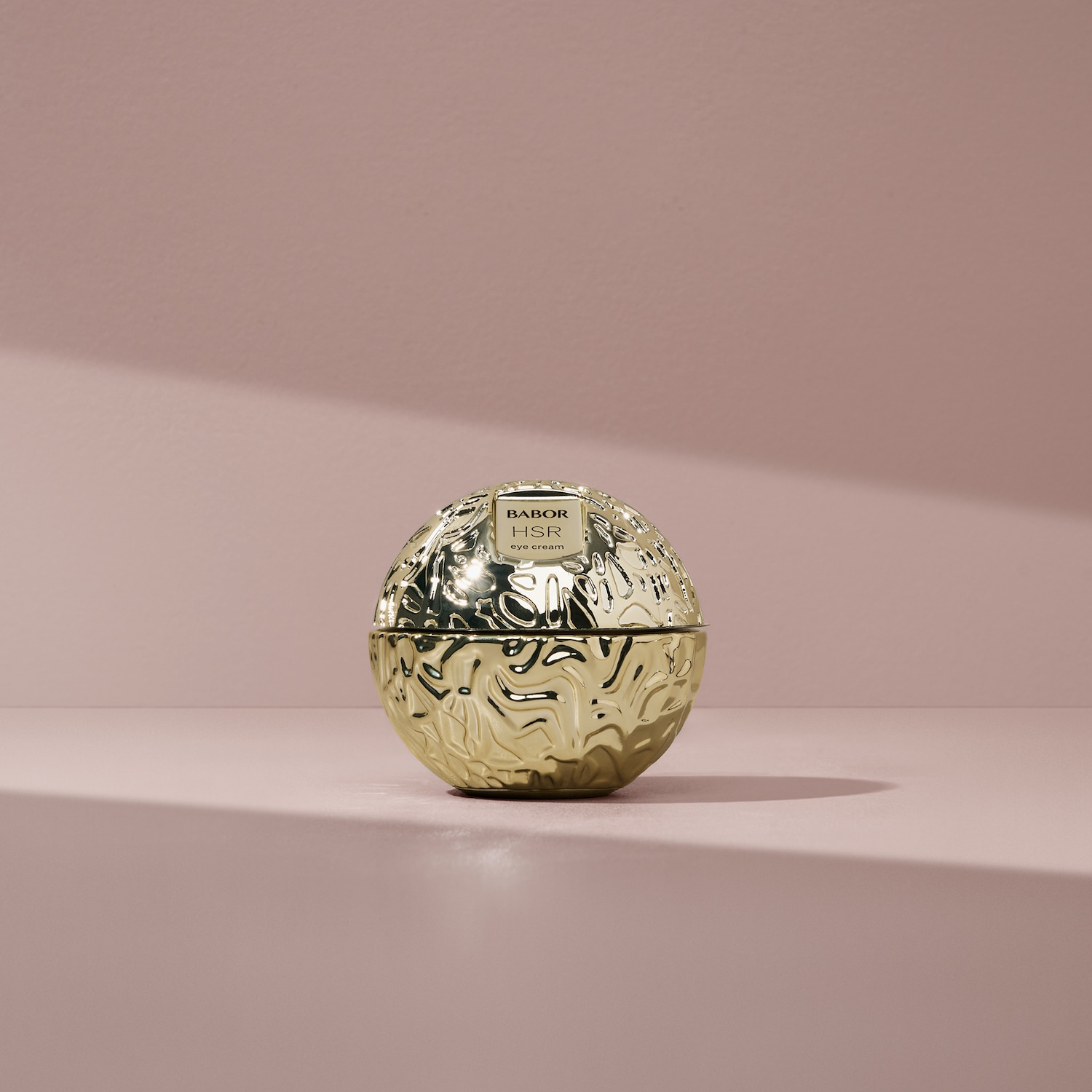 Złoty krem pod oczy Babor HSR w okrągłym opakowaniu o teksturowanej powierzchni, stojący na różowym tle, luksusowy produkt do pielęgnacji skóry.