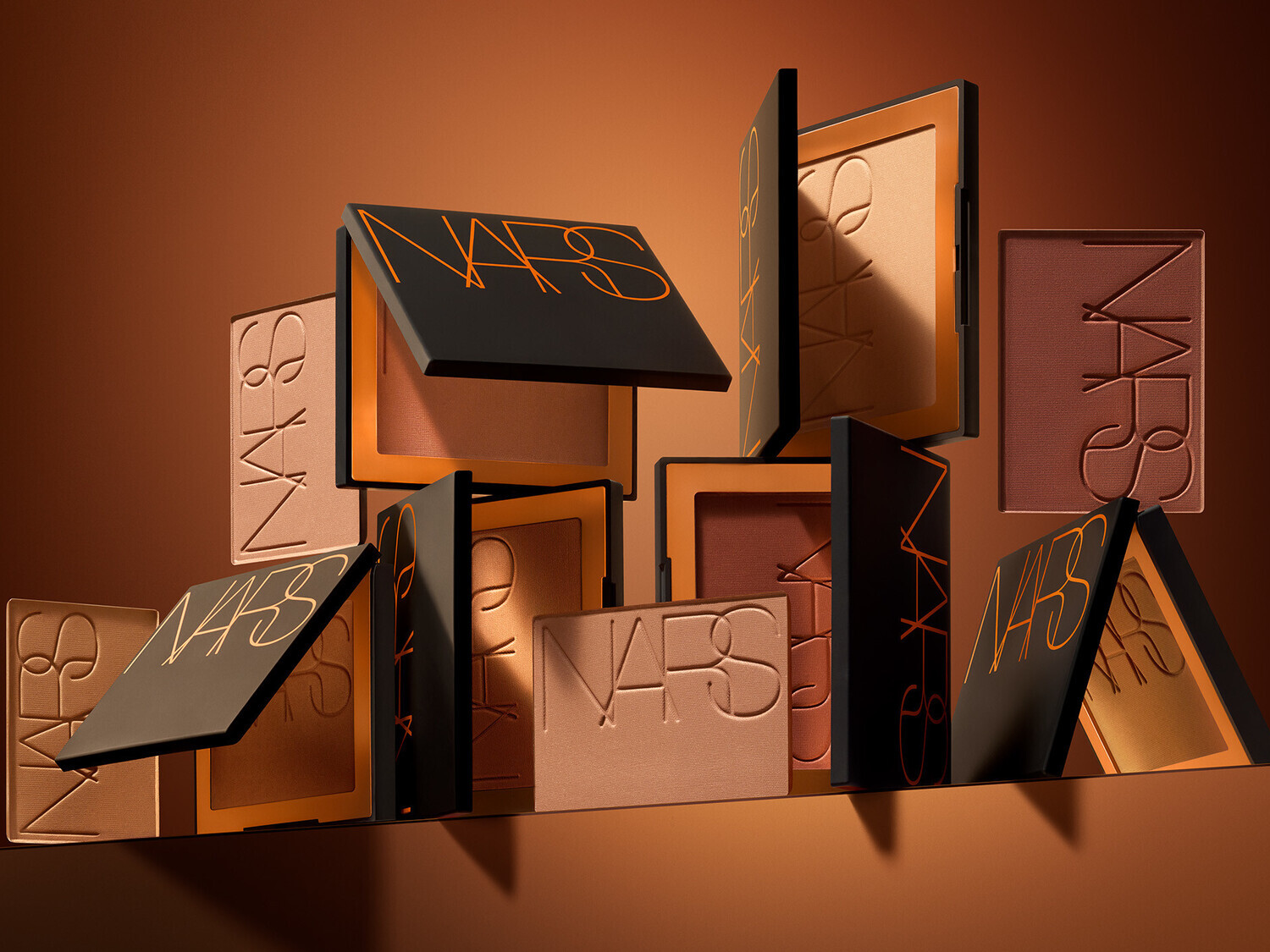 Una collezione di fard NARS in diverse tonalità di marrone, con confezioni nere e scritte arancioni, offre una gamma di opzioni per scolpire e illuminare il viso.