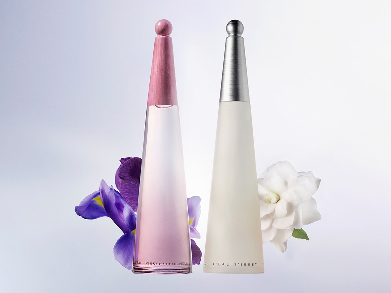 Dos frascos de perfume Issey Miyake se alzan con elegancia, uno en tonos rosados junto a una flor violeta, y el otro en blanco con una gardenia.
