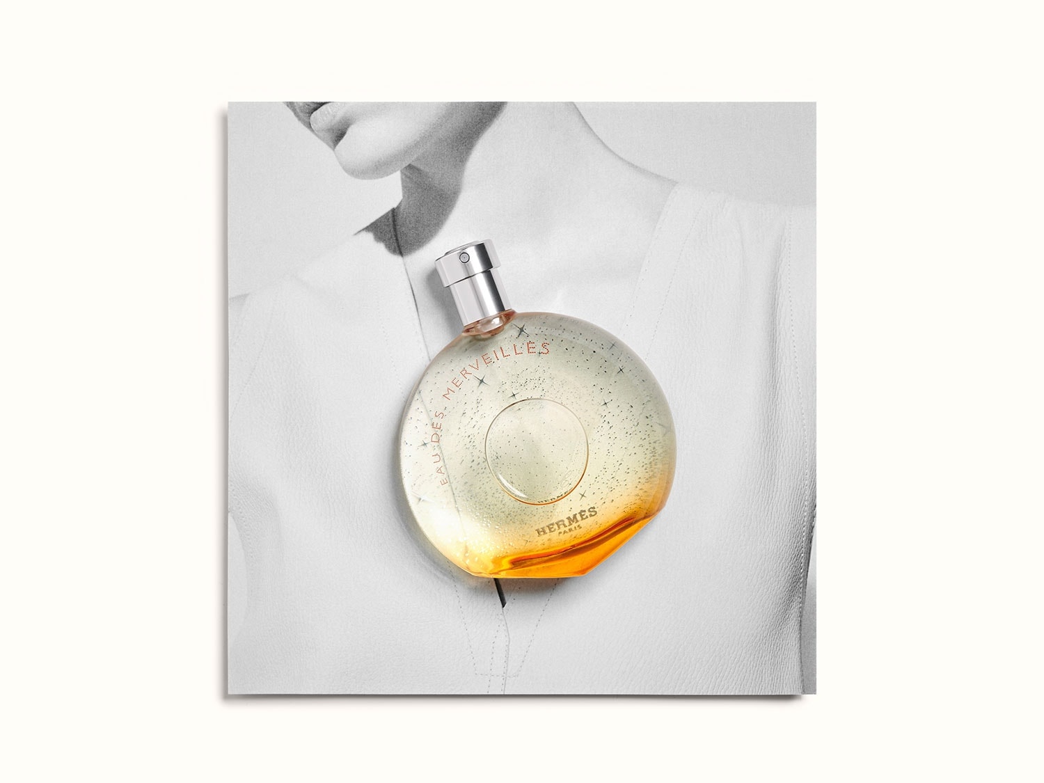 A kerek, narancssárga aljú, átlátszó Hermès Eau des Merveilles üveg egy nő fehér blúzán fekszik, ezüst kupakkal.
