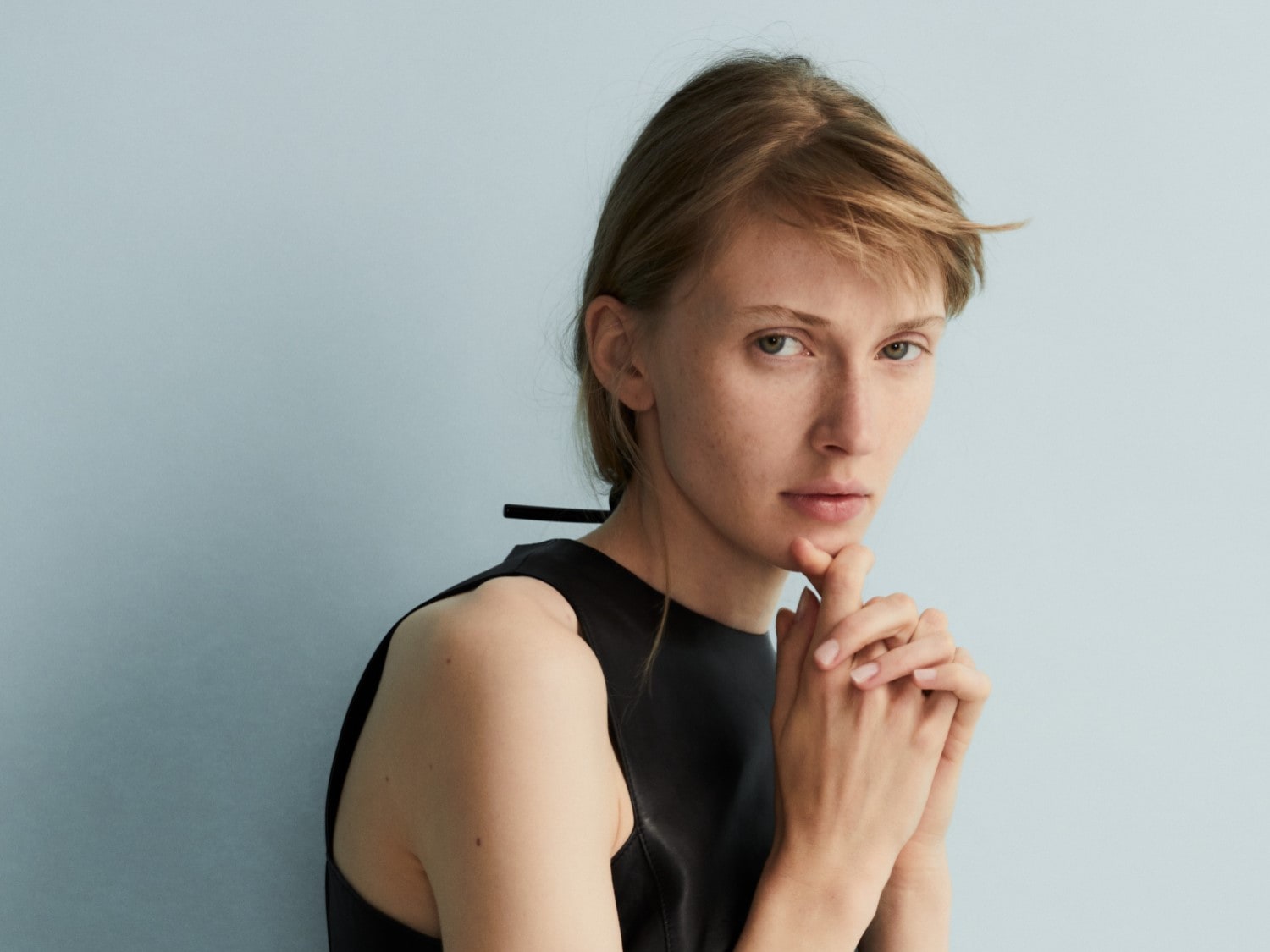 A világos bőrű, szőke hajú nő fekete, ujjatlan felsőt visel, és az állát támasztja a kezével, miközben a kamerába néz.