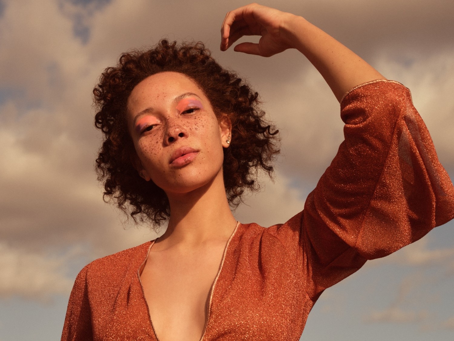 Portrét ženy s kučeravými vlasmi, pehami a oranžovými trblietavými šatami s farebnými očnými tieňmi pózuje s rukou nad hlavou na pozadí oblohy.