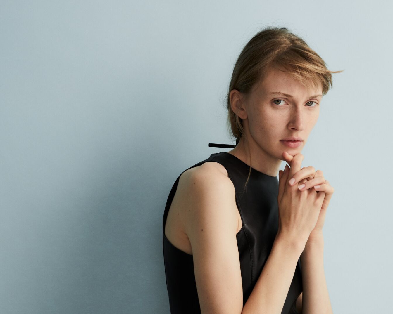 Portrét ženy s hnědými vlasy a v černých šatech bez rukávů, ruce má složené pod bradou a dívá se do objektivu.