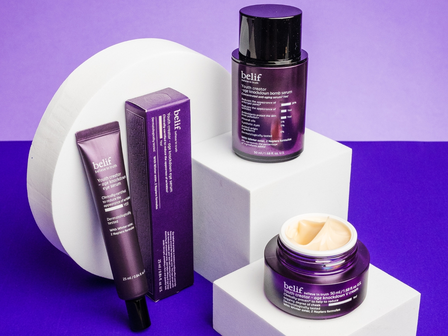 Na fioletowym tle znajdują się produkty Belif Youth Creator: serum do oczu, serum do twarzy i krem V, obiecujące redukcję zmarszczek i poprawę elastyczności skóry.