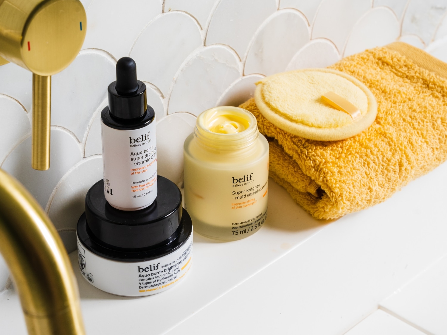 Na blacie łazienkowym stoją trzy produkty Belif: Aqua Bomb Super Drops z witaminą C w białej butelce z czarną zakraplaczem, Aqua Bomb Brightening z witaminą C i kwasem hialuronowym w białym słoiczku oraz Super Knights - multi vitamin sleep w żółtym słoiczku.