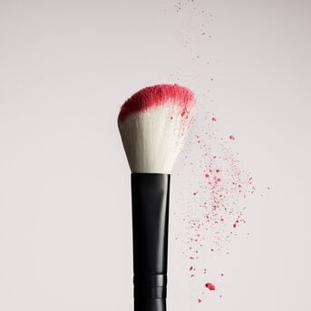 Detailní záběr štětce na make-up s černou rukojetí a bílými štětinami, které jsou na konci obarvené růžovým pudrem, s částečkami pudru vznášejícími se kolem.