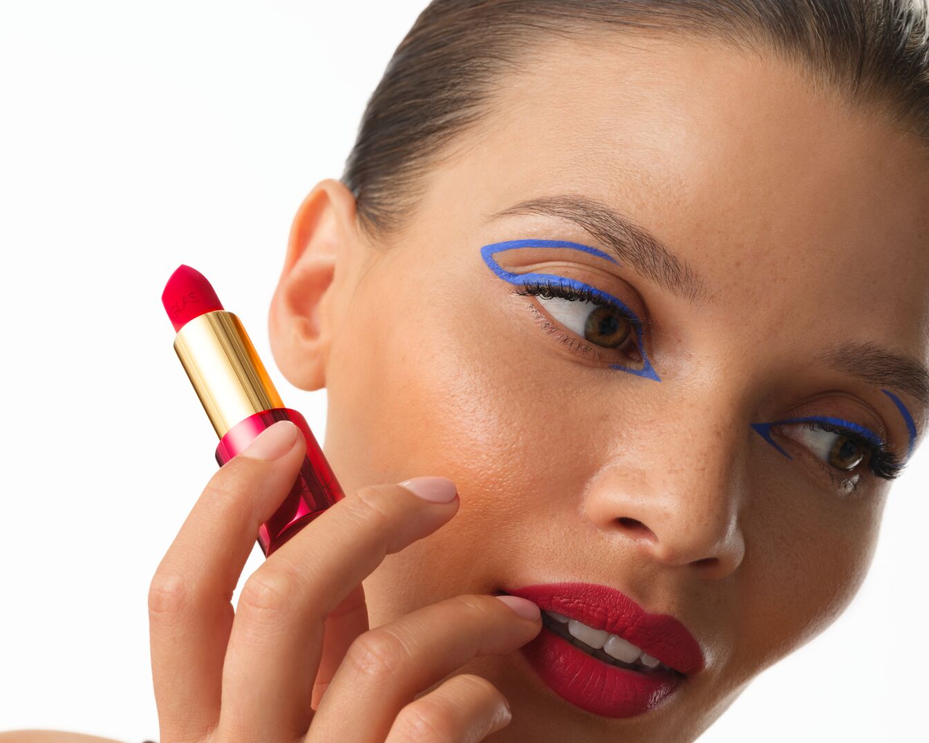 Detailní záběr na ženu s červenou rtěnkou a modrými očními linkami, která drží rtěnku ve zlatém obalu a dotýká se jí rty.