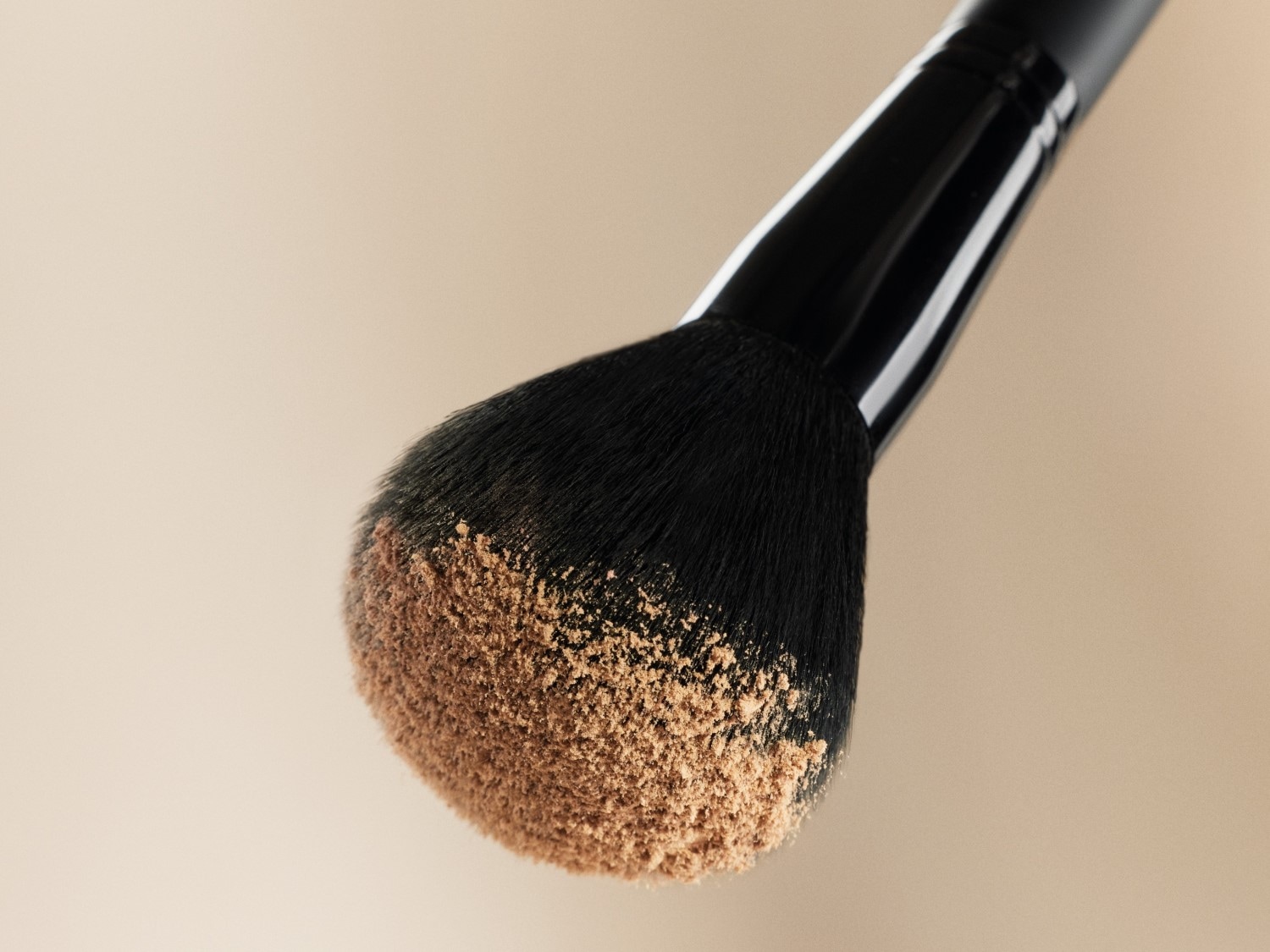 Detailní záběr na štětec na make-up s černou rukojetí a jemnými štětinami, které jsou lehce pokryté pudrovým make-upem.