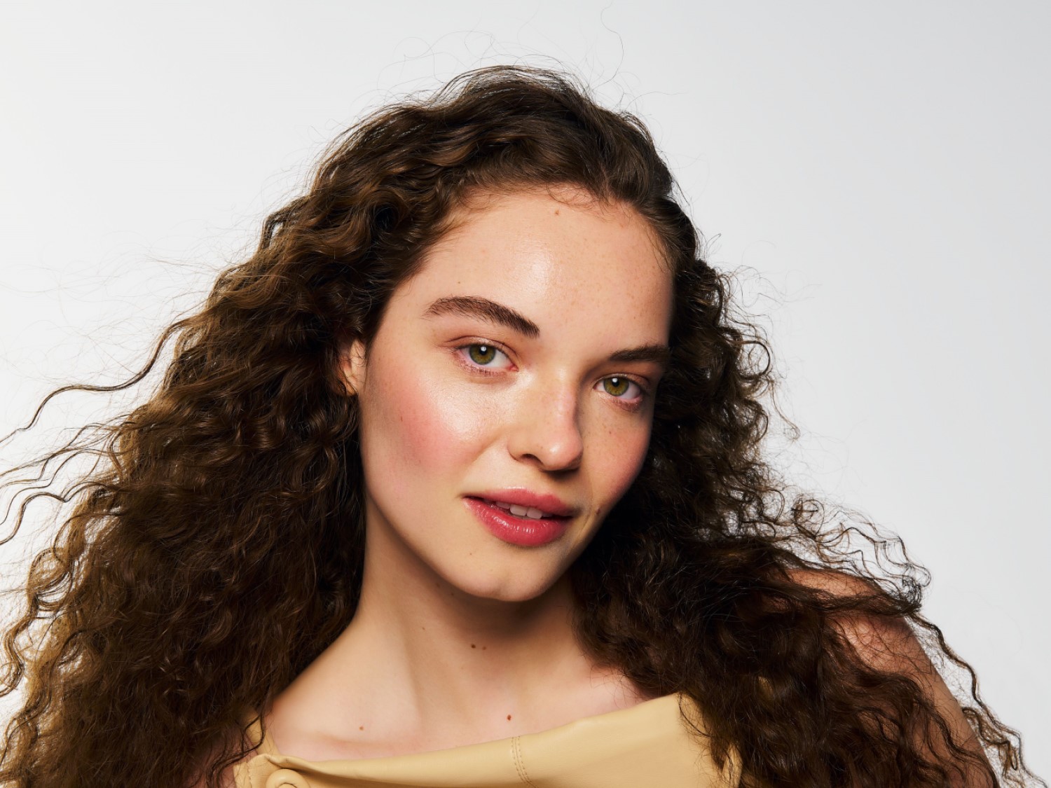 Portrét mladé ženy s vlnitými hnědými vlasy, světlou pletí a jemným růžovým make-upem, která se dívá do objektivu.