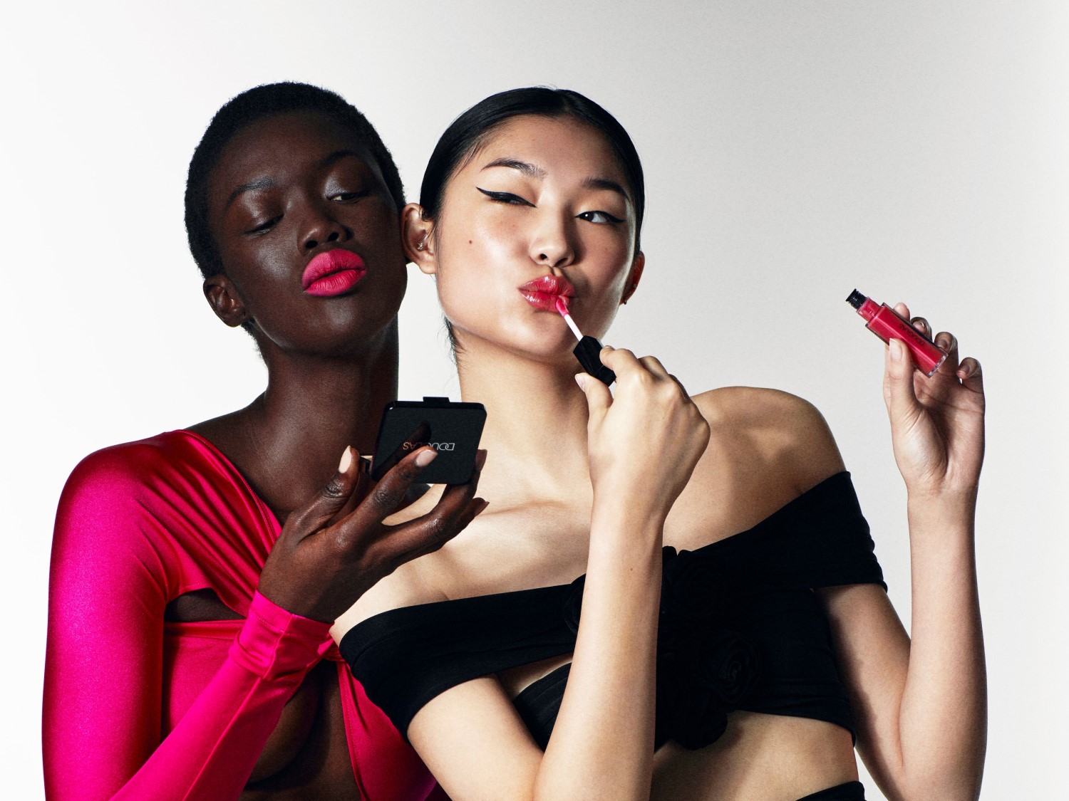 Két modell, az egyik élénk rózsaszín rúzst visel, és egy fekete kompaktot tart, míg a másik ajakfényt visz fel, és egy piros tubust tart.