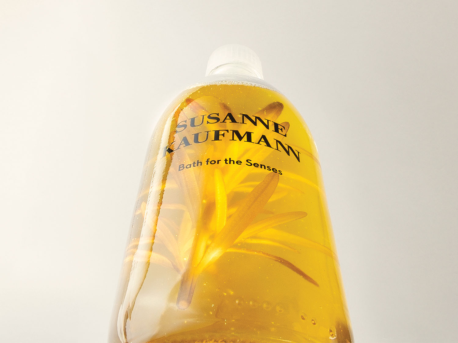 Die durchsichtige Flasche von Susanne Kaufmann enthält ein goldenes Badeöl mit dem Text „Bad für die Sinne“ und einem gelben Zweig.