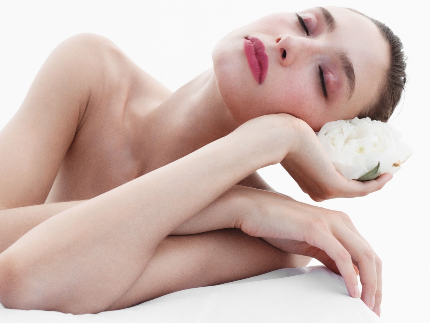 Eine Frau mit geschlossenen Augen ruht sich aus, ihr Kopf liegt auf einer weißen Blüte, die sie in der Hand hält, und betont so die Ruhe und Reinheit des Moments.