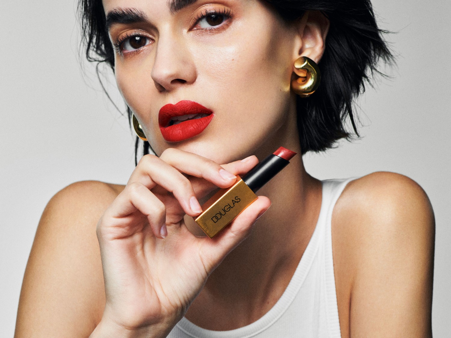Eine Frau mit roten Lippen hält einen Douglas Lippenstift in der Hand, dessen goldene Verpackung elegant hervorsticht.