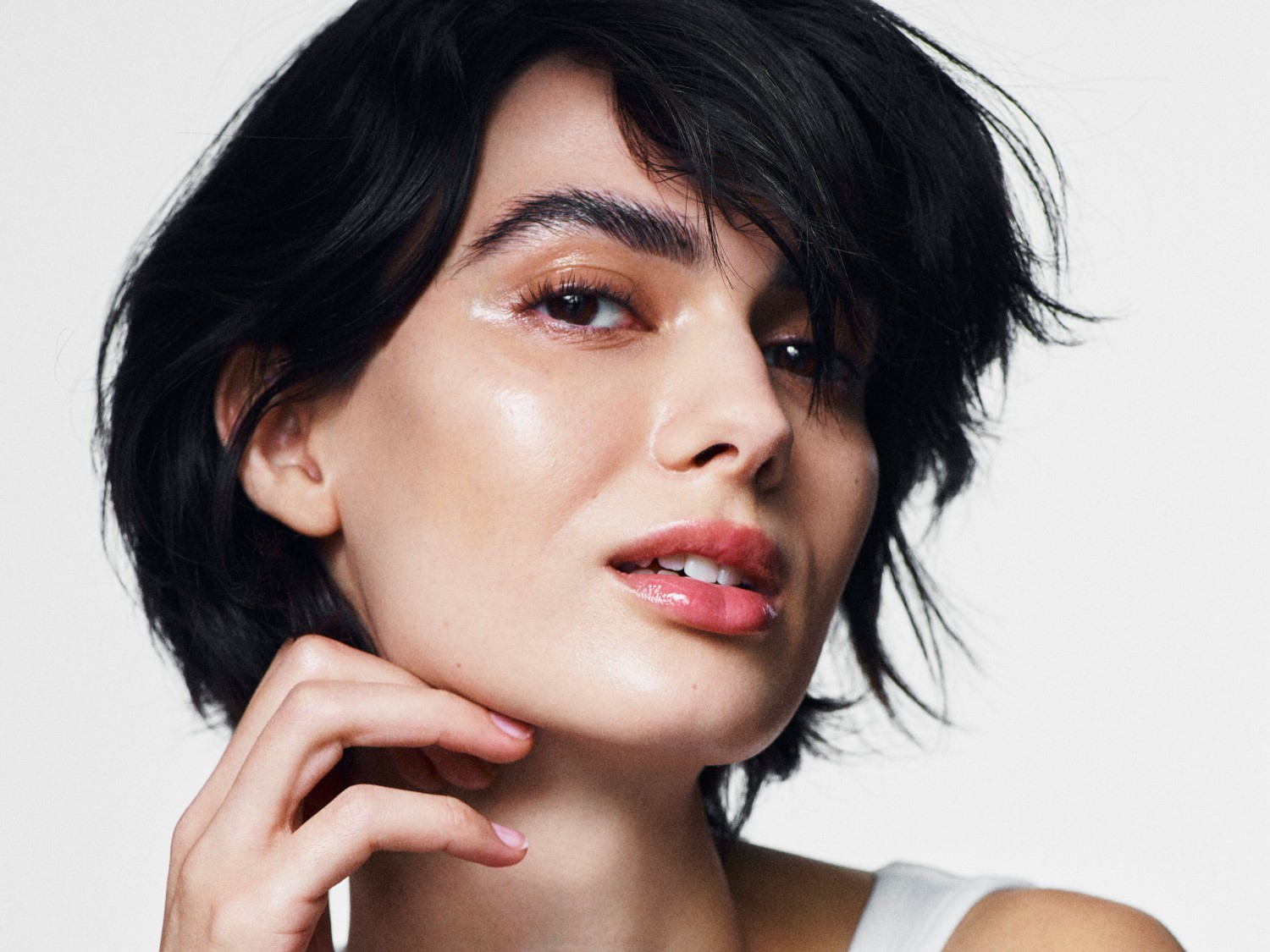 Nahaufnahme einer Frau mit glänzender Haut, dunklen, gewellten Haaren und einem natürlichen Make-up, die ihre Wange berührt.