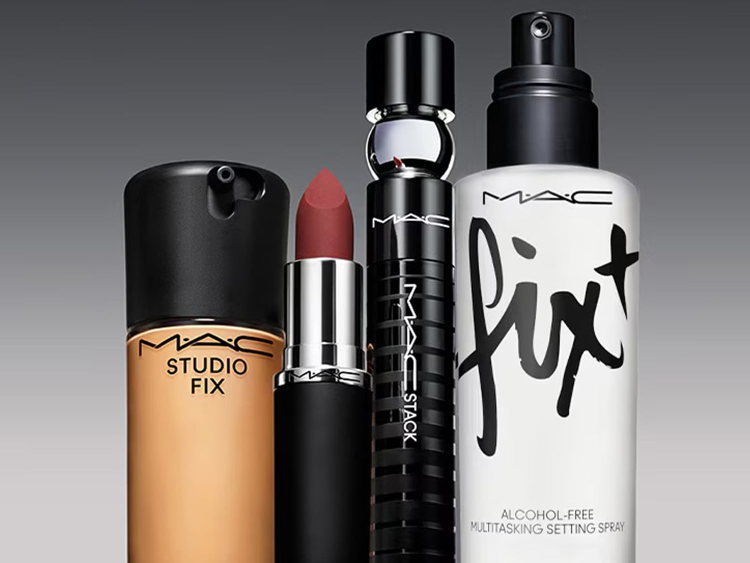 Čtyři produkty MAC Cosmetics jsou zobrazeny na šedém pozadí: podkladová báze Studio Fix v béžové lahvičce s černým uzávěrem, matná rtěnka v odstínu cihlově červené, řasenka MAC Stack v černé tubě a fixační sprej Fix+ Stay Over v bílé lahvičce s černým rozprašovačem.