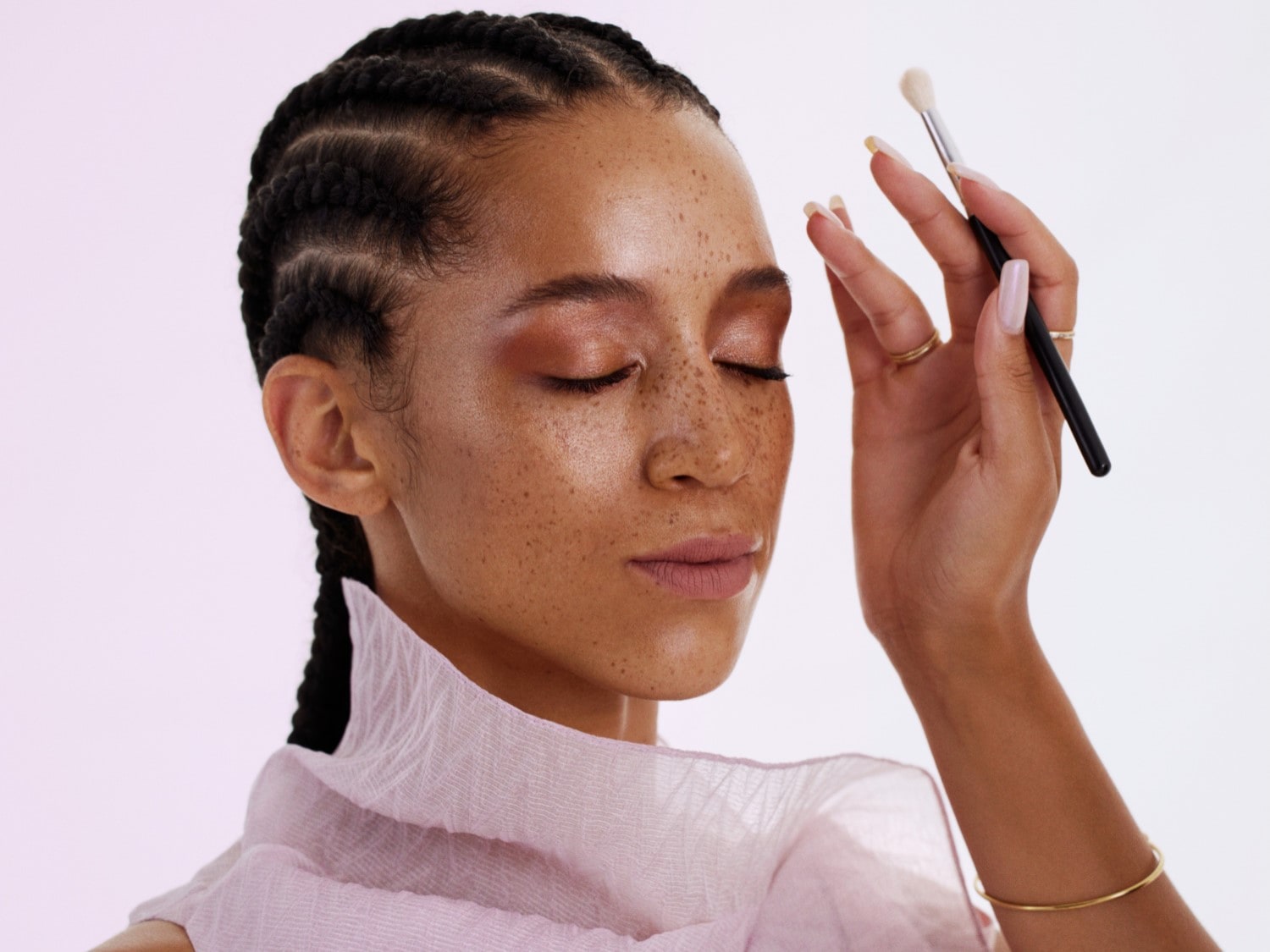 Žena s copánky a pihami si nanáší štětcem oční stíny, má zavřené oči a na sobě světle růžový šátek.