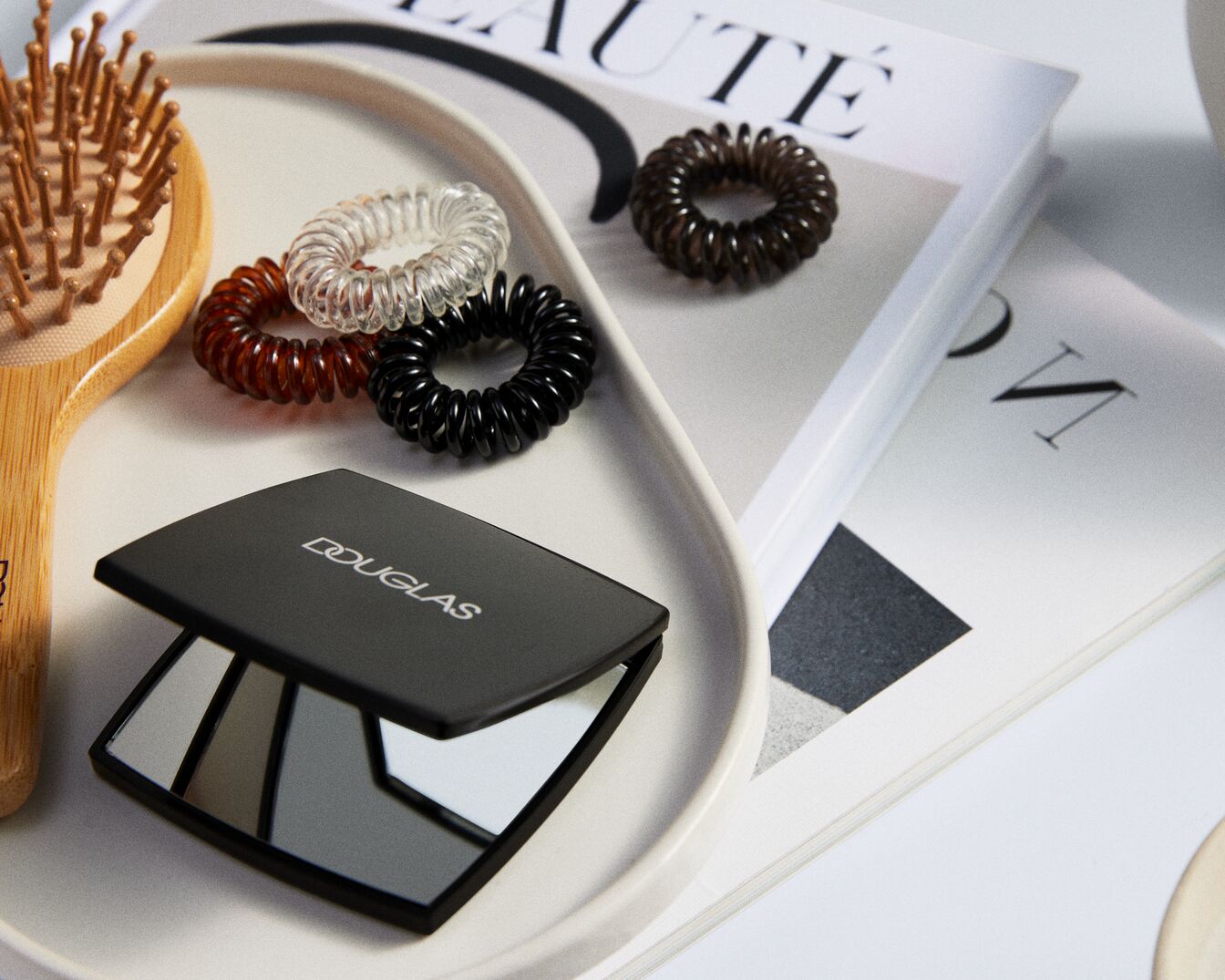 Een make-upspiegel van Douglas ligt op een wit dienblad, samen met een houten haarborstel en spiraalvormige haarelastiekjes in bruin, zwart en transparant.