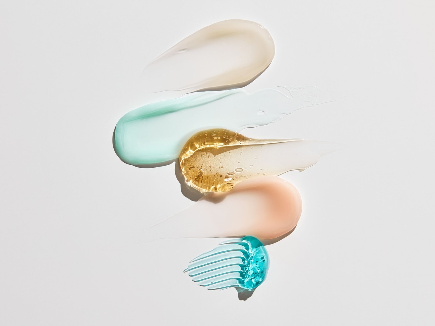 Pět tahů různých kosmetických produktů, včetně bílého krému, tyrkysového gelu, zlatavého séra, broskvové emulze a modrého gelu, které ukazují textury a barvy.