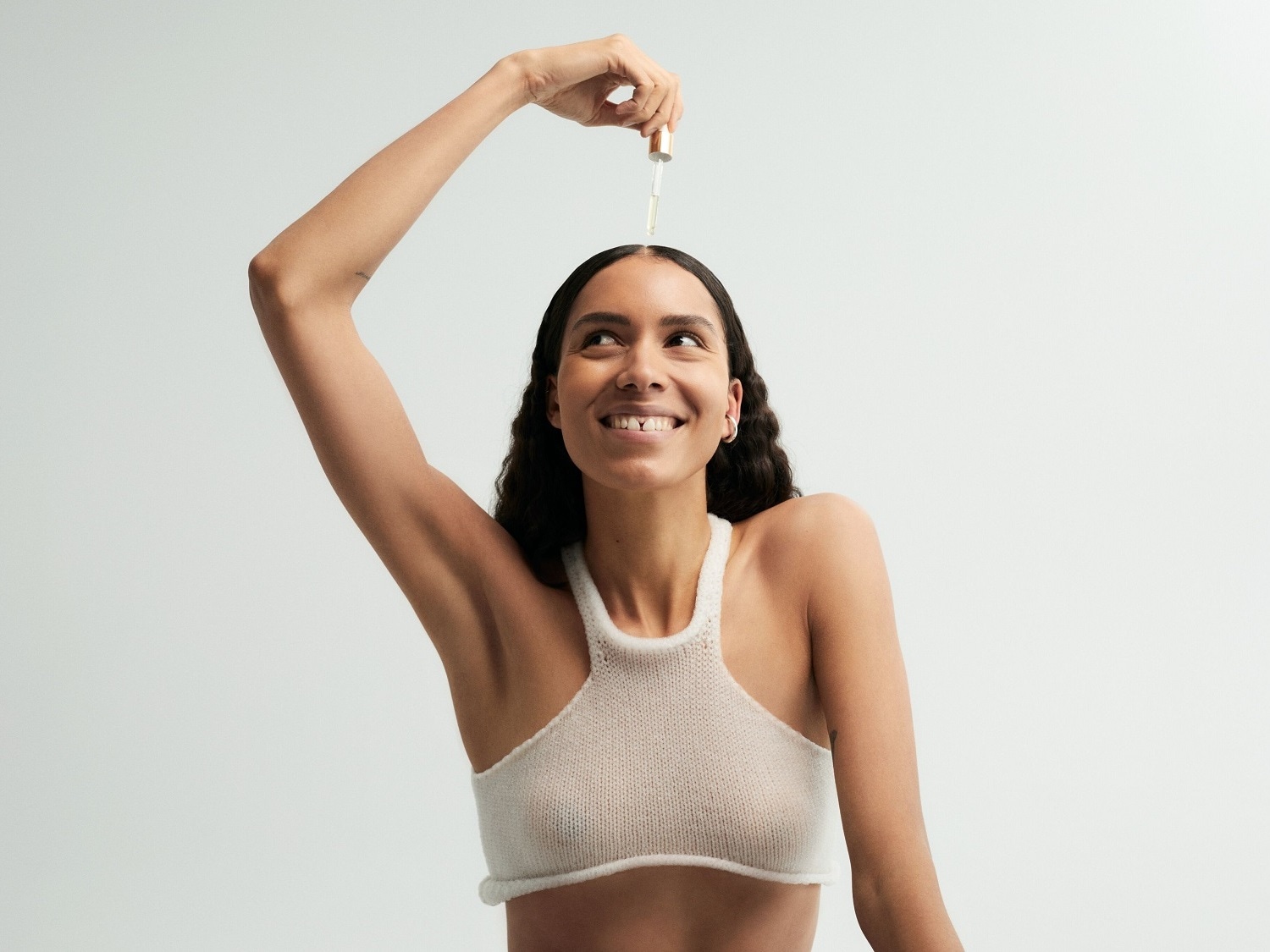 Švytinti moteris su šviesiu viršumi, pakėlusi ranką su serumo buteliuku, lašinančiu ant jos plaukų, žiūri aukštyn su šypsena.