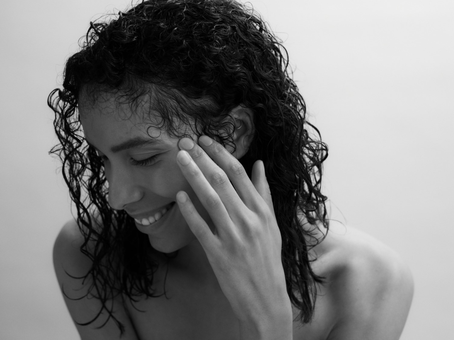 Černobílý portrét usmívající se ženy s mokrými kudrnatými vlasy, která se dotýká tváře.