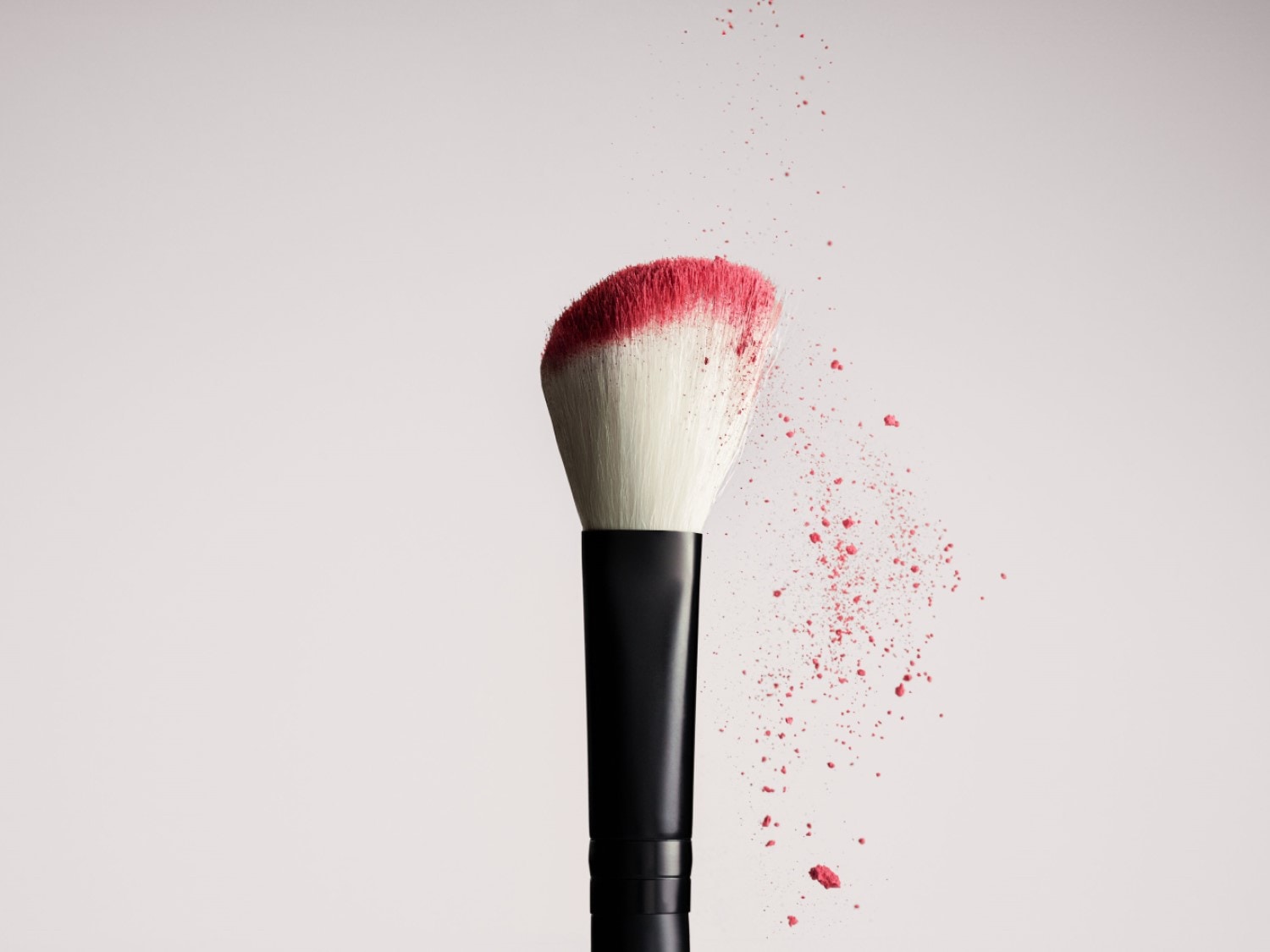 Detailný záber štetca na make-up s čiernou rúčkou a bielymi štetinami, ktoré sú na konci zafarbené do ružova, okolo poletujú čiastočky ružového púdru.