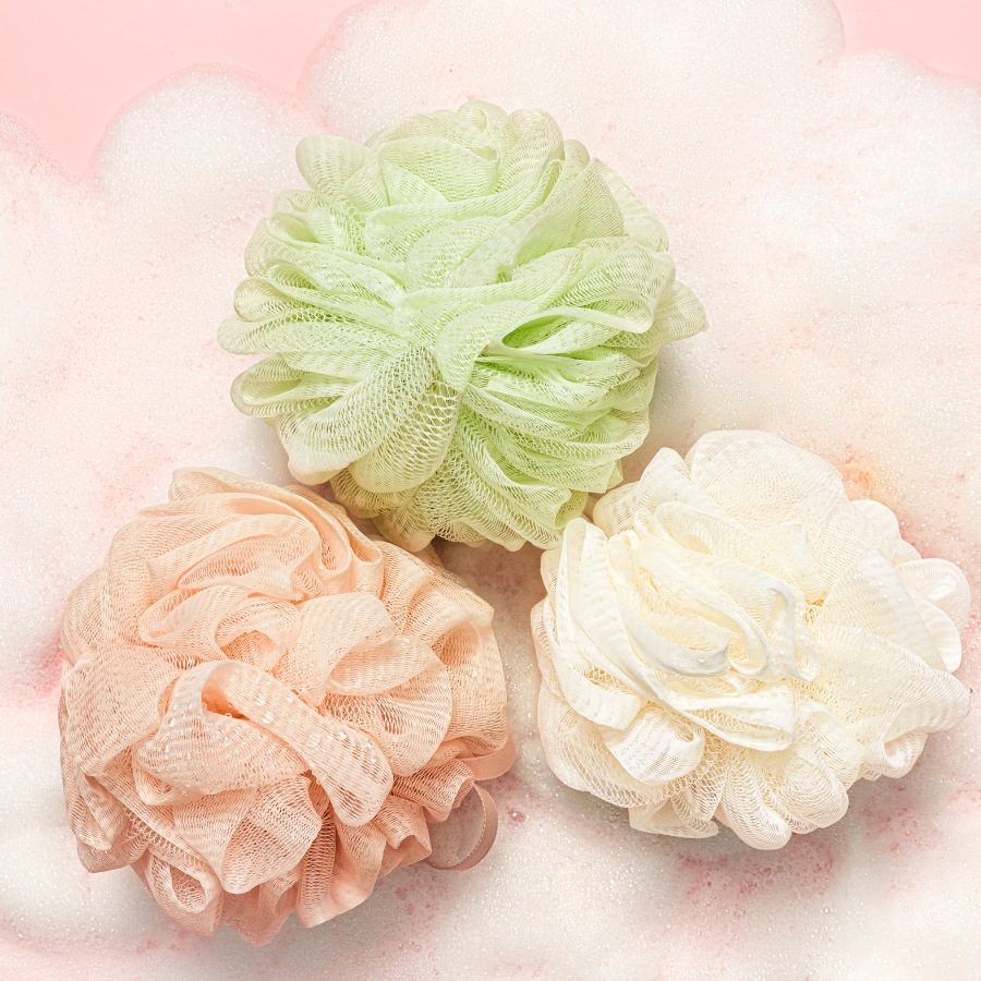 Trzy kolorowe myjki do ciała: zielona, brzoskwiniowa i kremowa, leżą na tle różowej powierzchni pokrytej pianą.