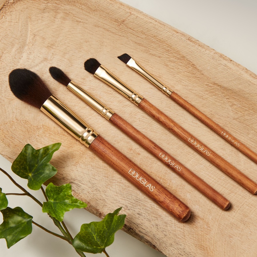 Cztery pędzle do makijażu Douglas z drewnianymi rączkami i złotymi skuwkami, ułożone na drewnianej desce obok zielonych liści.