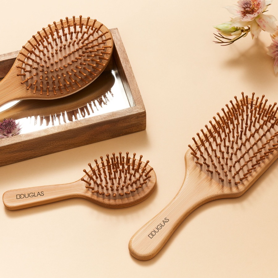 Trzy bambusowe szczotki do włosów Douglas leżące na beżowym tle, obok kwiatów.