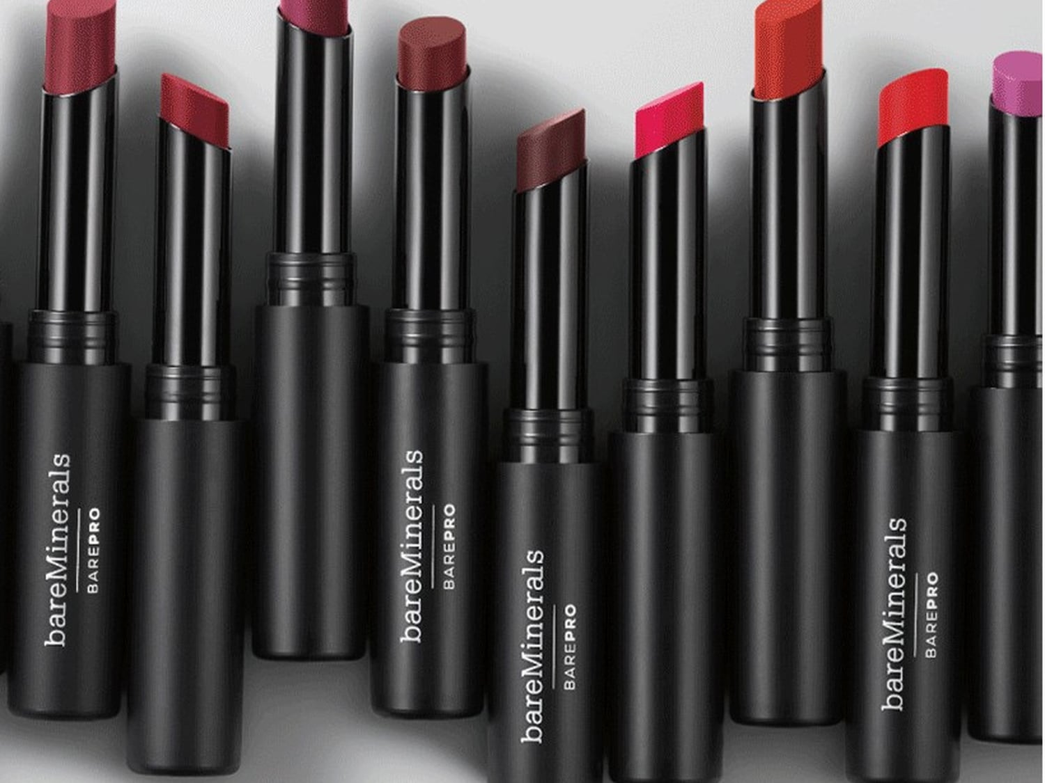Różnokolorowe szminki bareMinerals w czarnych, smukłych opakowaniach, ustawione obok siebie, prezentują szeroką gamę odcieni.