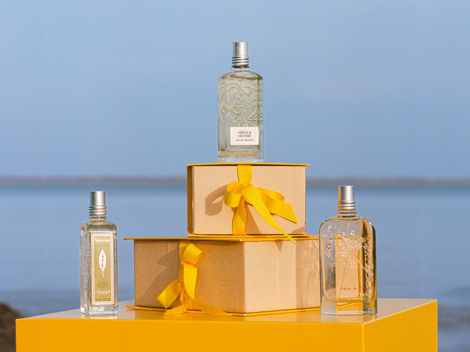 Na żółtym podeście stoją dwa pudełka z żółtymi wstążkami, na których umieszczono trzy flakony perfum L'Occitane o różnych zapachach.