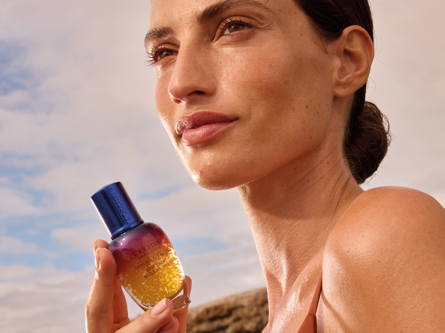 Kobieta trzyma w dłoni buteleczkę olejku do twarzy L'Occitane Immortelle Reset Nuit Huile-En-Serum z niebieską nakrętką, na tle nieba.