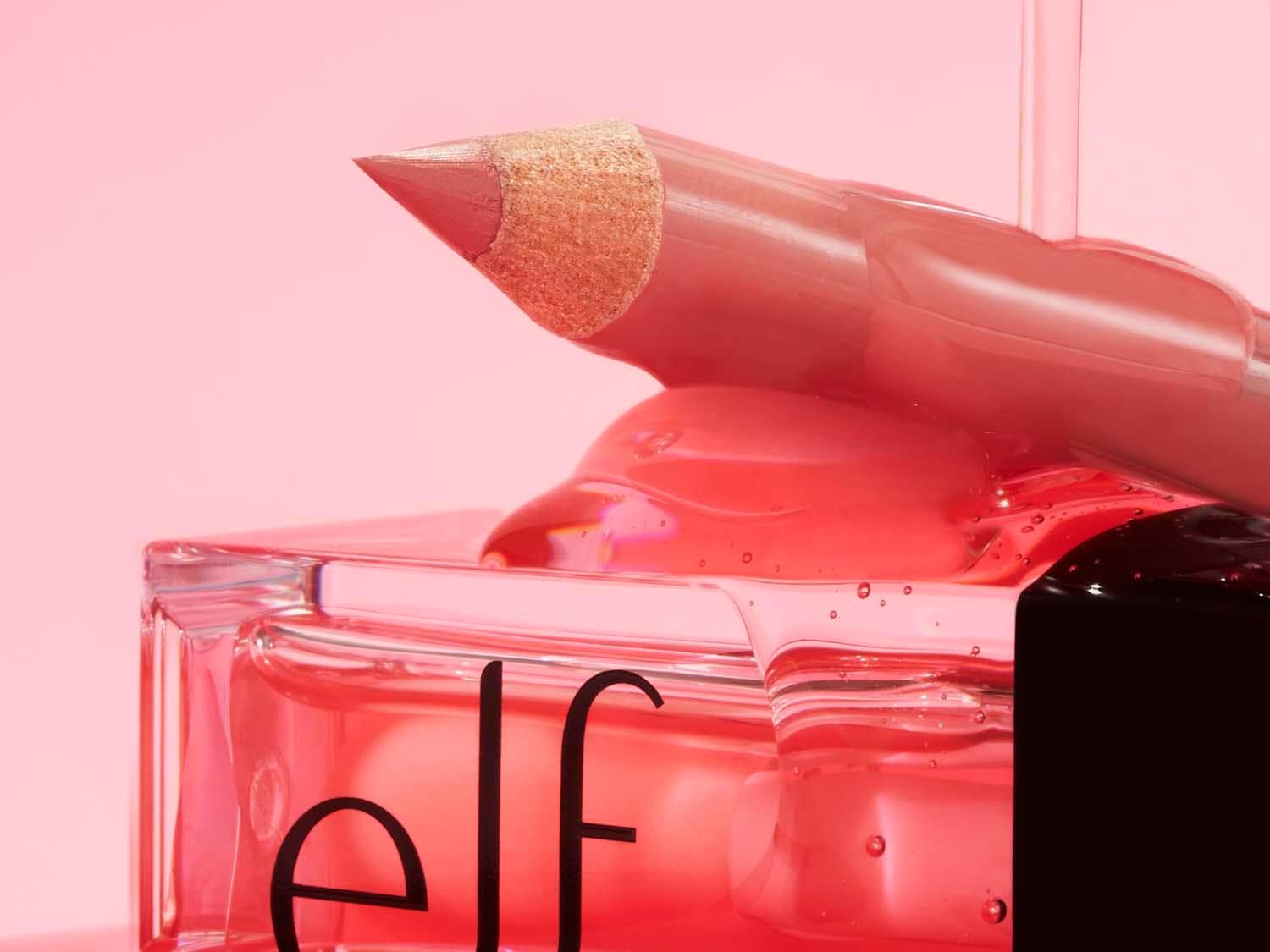 Close-up van een transparante fles met roze vloeistof, een lippotlood en een roze lipgloss, allemaal van e.l.f. Cosmetics, tegen een roze achtergrond.