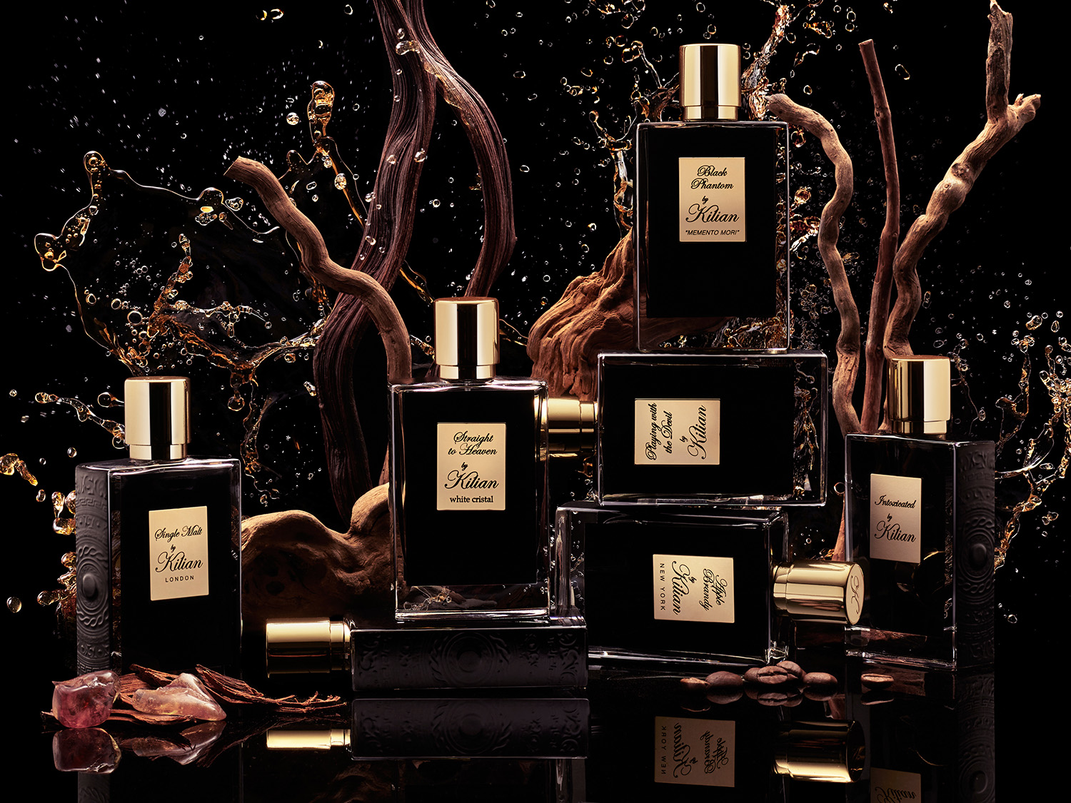 Fekete háttér előtt Kilian parfümök gyűjteménye látható, arany kupakokkal és feliratokkal, faágakkal és kávébabokkal díszítve.