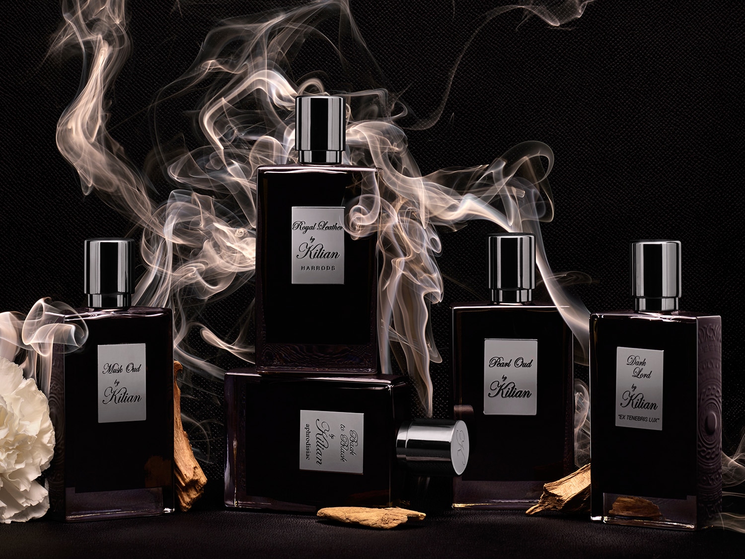 Kilian parfümök láthatók különböző illatokban, mint például a Musk Oud, a Royal Leather és a Dark Lord, füstös háttérrel és fadarabokkal díszítve.