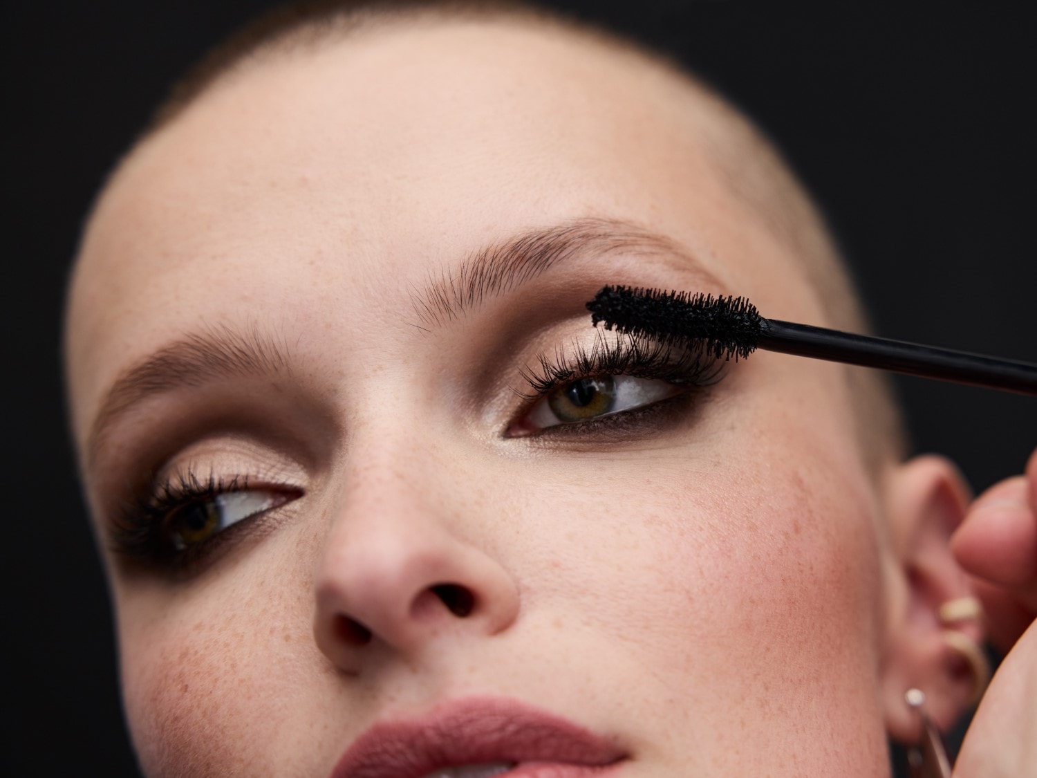 Detailný záber ženy s krátkymi vlasmi, ktorá si nanáša čiernu maskaru na horné riasy, aby zvýraznila svoje zelené oči.